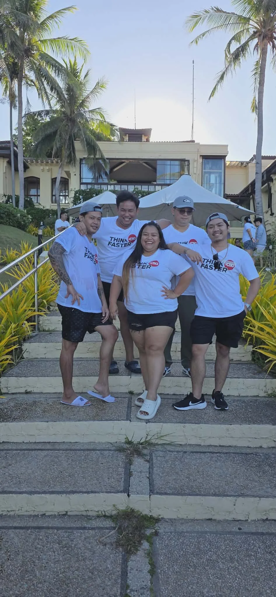 Group of five smiling people standing on steps outdoors, wearing matching white 'Think Faster' t-shirts, with a building’s porch, potted plants, and palm trees in the background.