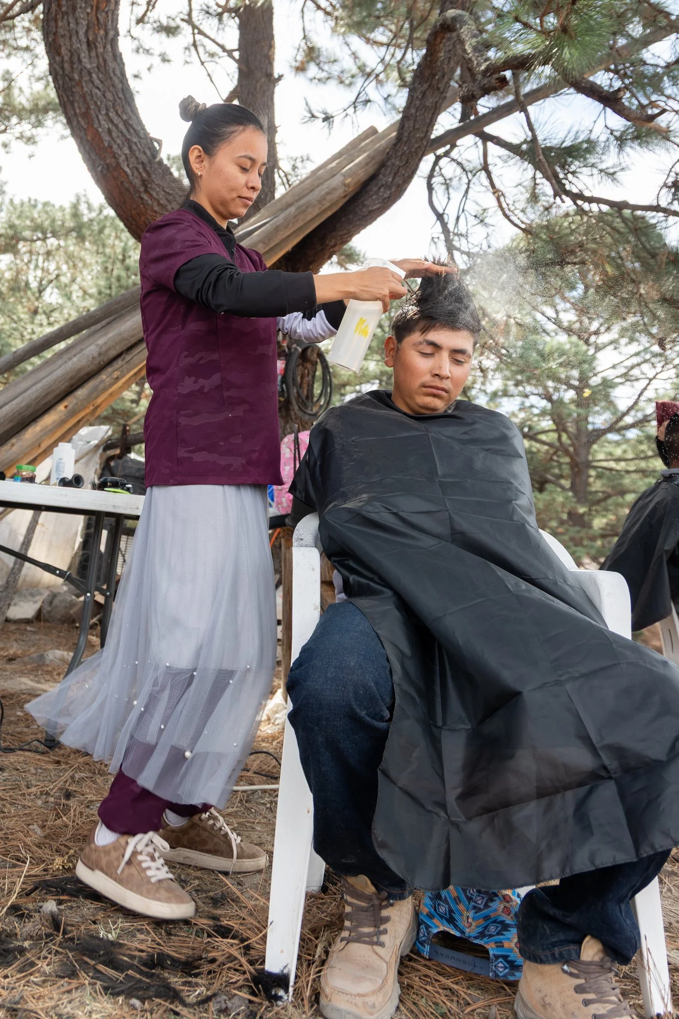 A woman giving a haircut outdoors under a tree to a man sitting in a chair with a black cape. The woman is holding a spray bottle and the man has a neutral expression. There are trees and outdoor supplies - Sasha at REDFISH | Creative Studio
