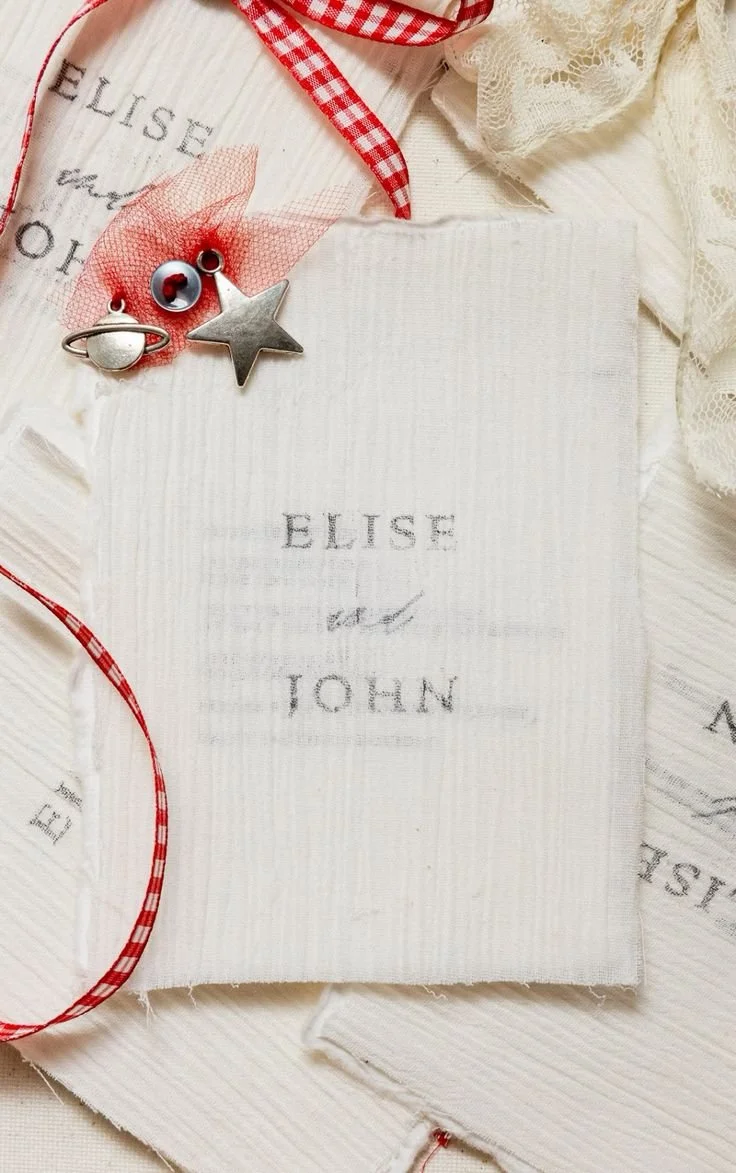 Textiles with embroidered names 'Elise and John', red checkered ribbons, lace fabric, and metal charms including a star, a ring, and a red eye symbol, arranged decoratively.