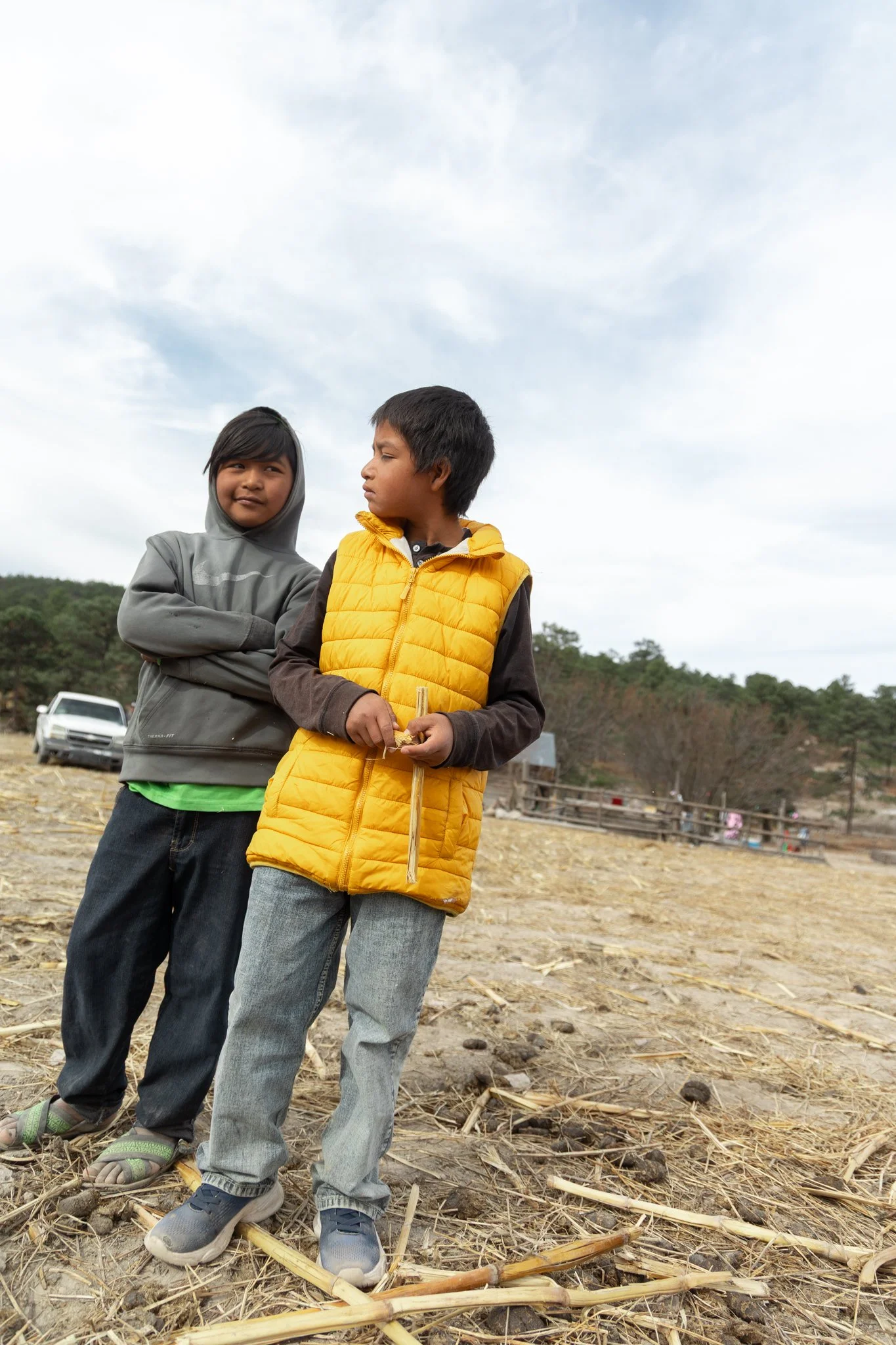 Two children standing outdoors in a field with cloudy sky, one in a gray hoodie and the other in a yellow vest, holding a stick, with a car and trees in the background - Sasha at REDFISH | Creative Studio