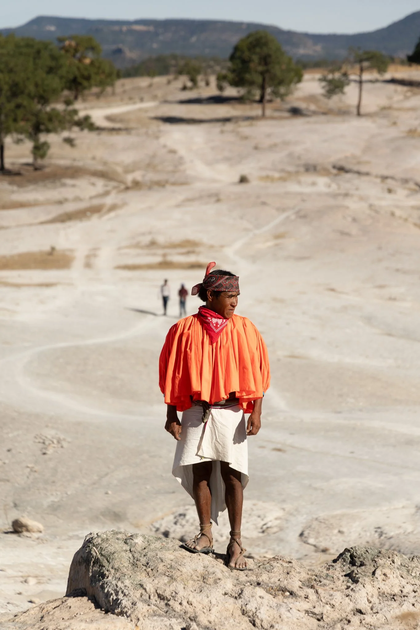 A person wearing traditional attire, including an orange cape, white skirt, and sandals, standing on a large rock in a barren desert landscape with a few trees and distant mountains.