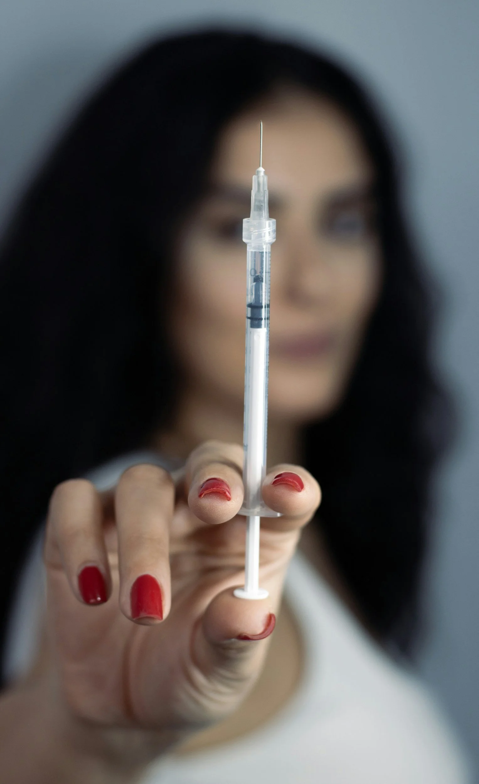 A woman with dark hair and red nail polish holding a syringe filled with liquid, positioning it towards the camera. The focus is on the syringe, with the woman's face blurred in the background.