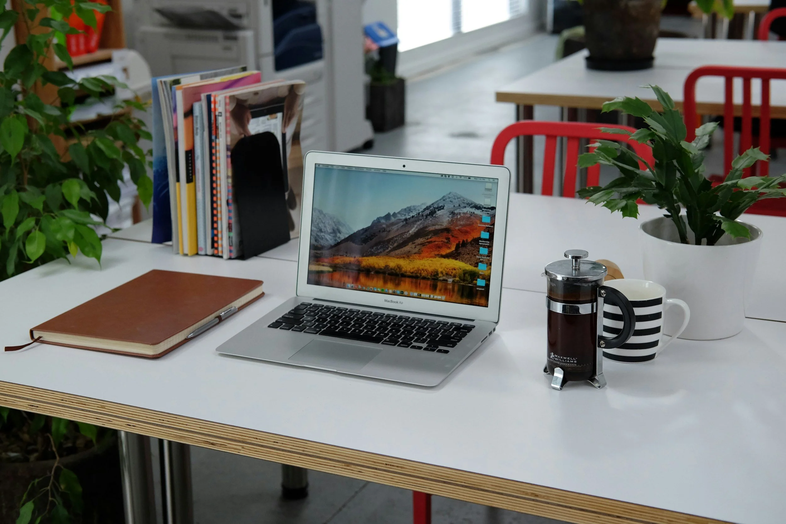 A workspace with a laptop, a closed brown notebook, a French press, a striped mug, a potted plant, and a stack of books and magazines on a white table in an office or library setting.