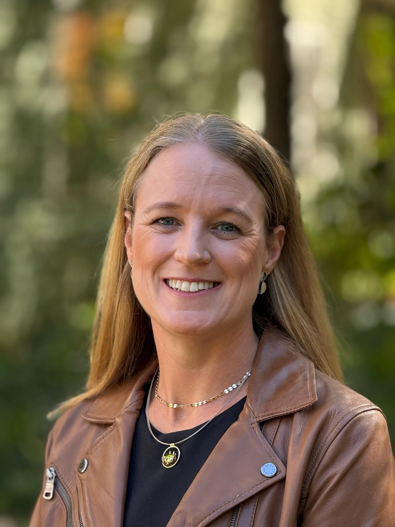 A smiling woman standing outdoors with a blurred green and brown forest background. She has long light brown hair, light blue eyes, and is wearing a brown leather jacket, a black top, and layered necklaces.