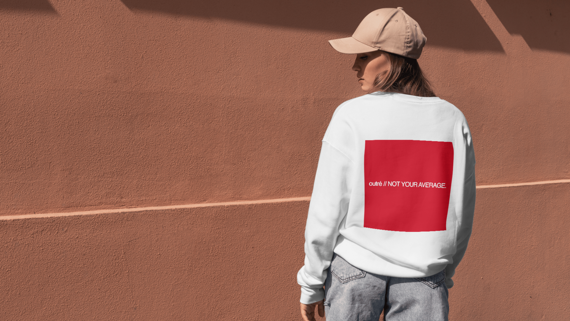 A woman wears a white sweater with a red square on the back that says "outré // Not Your Average".