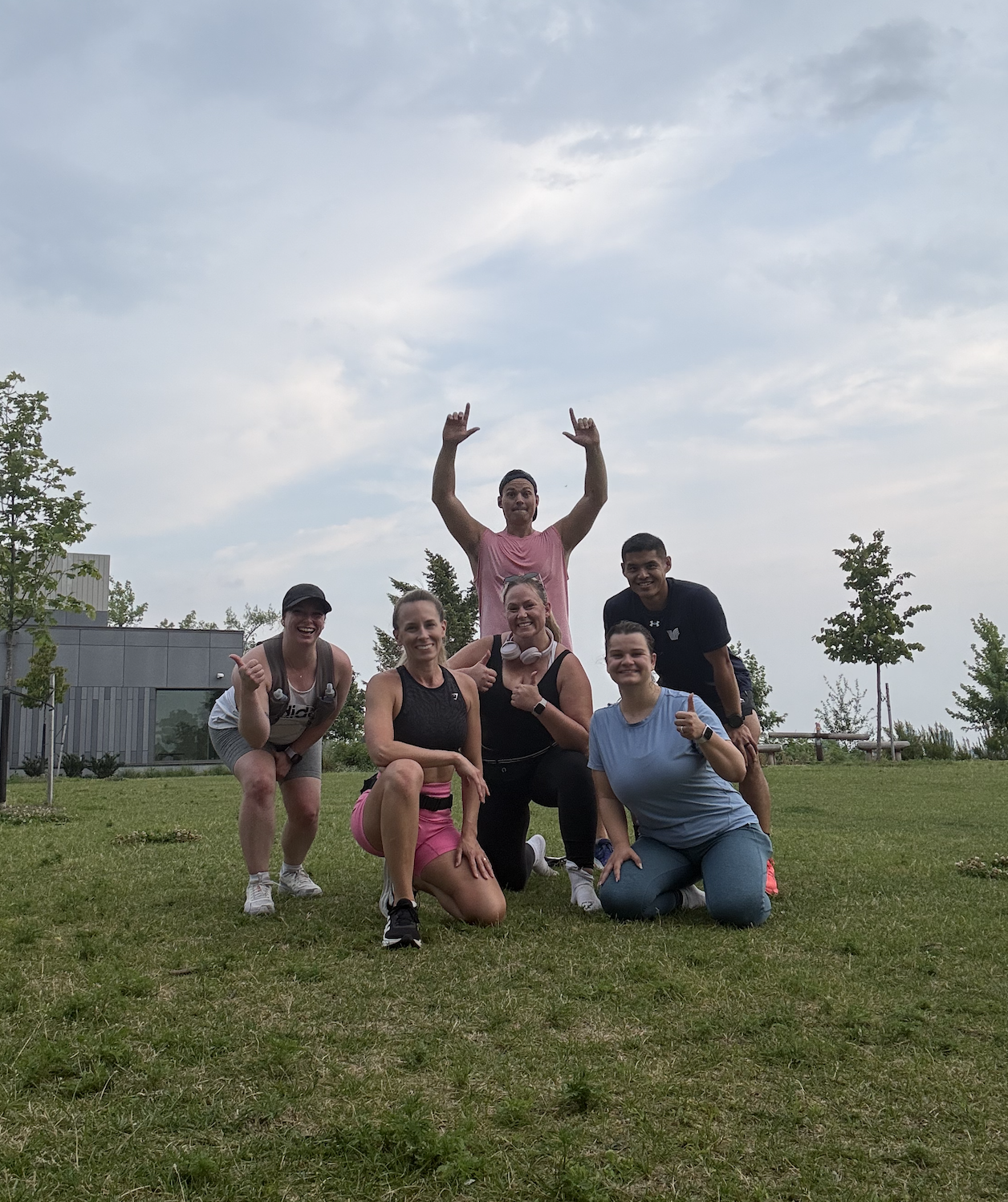 Group of six people outdoors in a grassy area, posing together and smiling. Some are kneeling or squatting, with arms around each other, celebrating or having fun, with cloudy sky and modern building in the background.