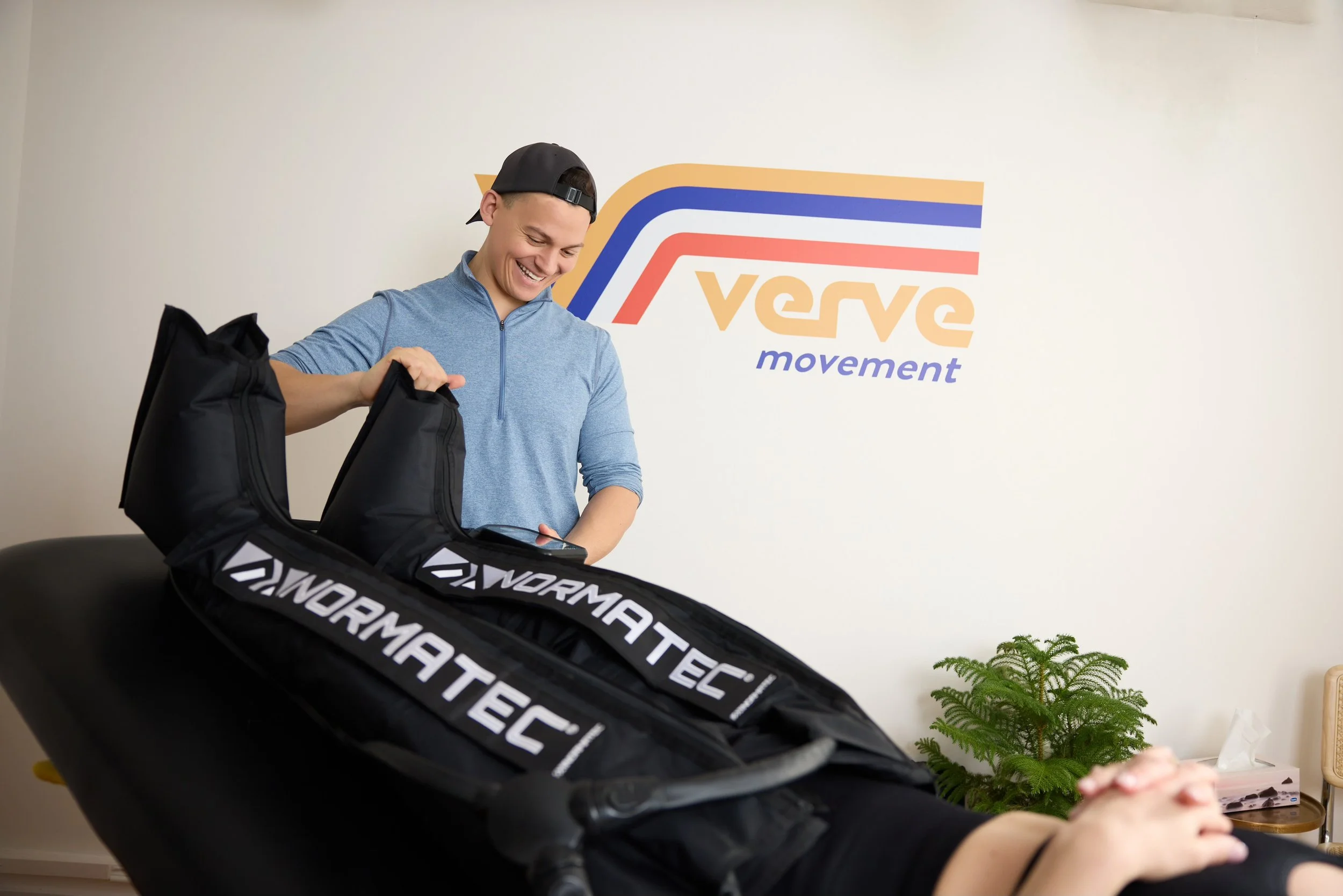 Person using Normatec compression therapy on a client in a room with "Verve Movement" signage and a plant in the background.