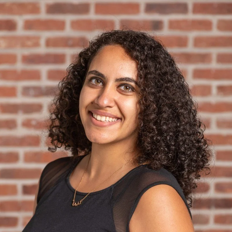 A woman with curly brown hair smiling at the camera, wearing a black sleeveless top and a gold necklace, with a brick wall background.
