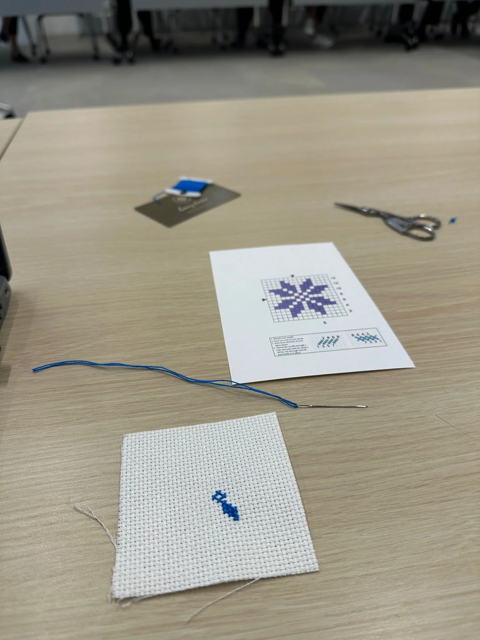 A cross-stitch craft project on a light wood desk, with a small piece of white fabric stitched with a blue pattern, a needle threaded with blue thread, a pair of scissors, and a printed pattern guide.