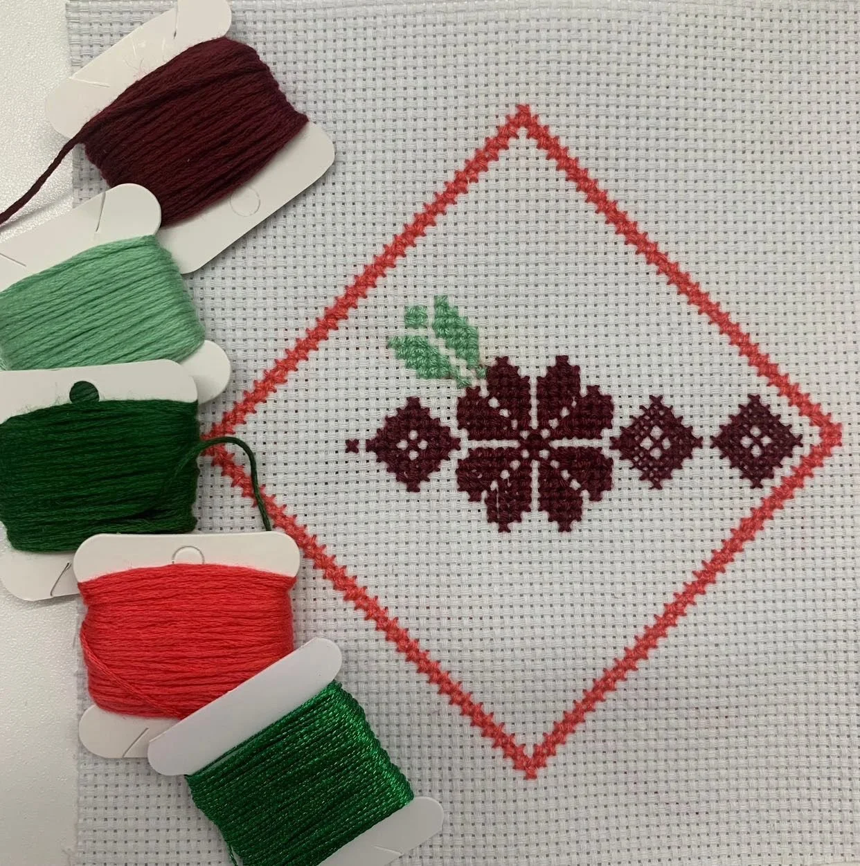 Cross-stitch embroidery of a geometric floral pattern within a red diamond border on white fabric, with five spools of embroidery thread in shades of red and green to the left.