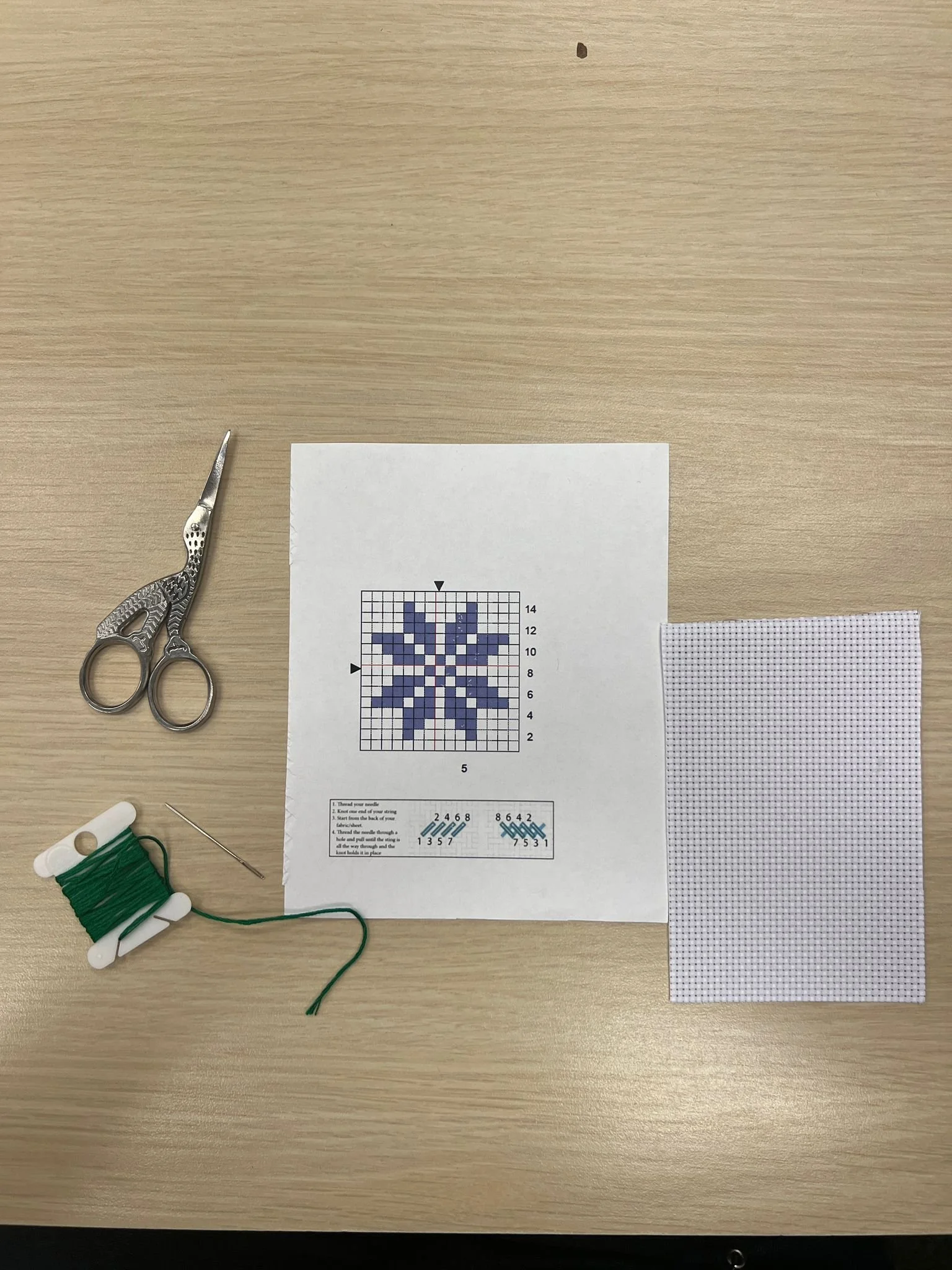 Cross-stitch embroidery kit with a pattern, a spool of green thread, a needle, a pair of scissors, and cross-stitch fabric on a wooden table.