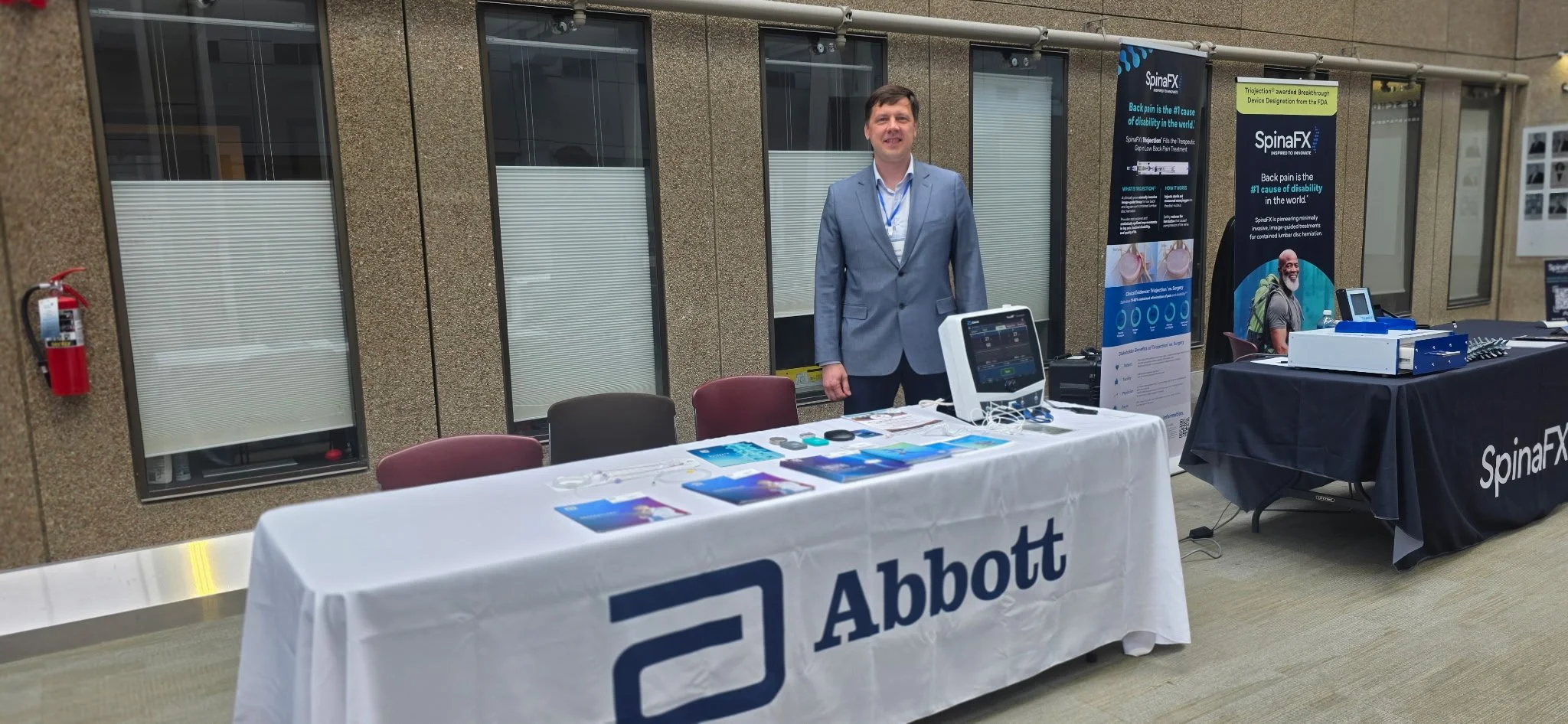 A man in a gray suit standing behind a table with Abbott branding at an outdoor event. The table displays pamphlets and medical devices. There are two banners about SpinaFX therapy and a digital display device on the table. A fire extinguisher is mou