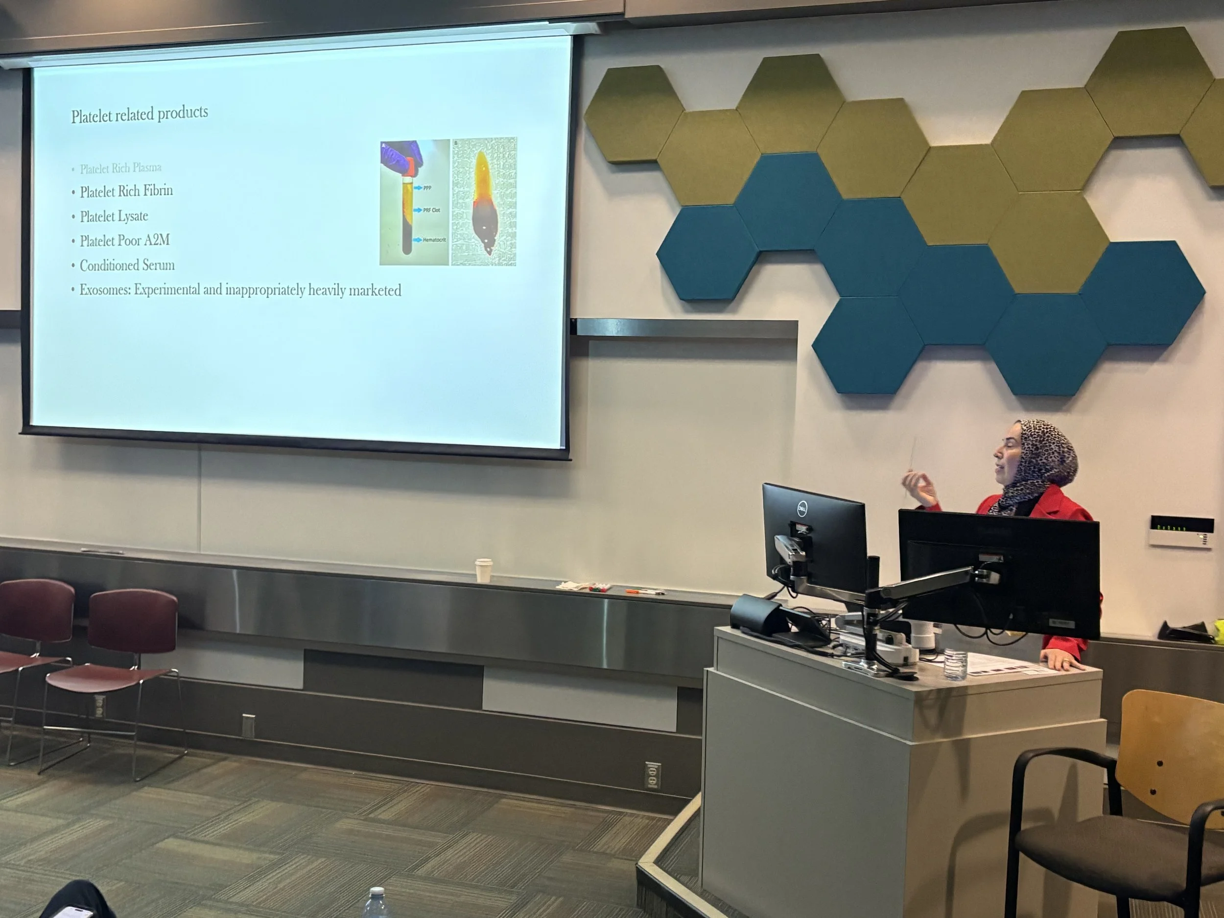 A woman wearing a hijab and red jacket is standing at a podium with a computer and microphone, giving a presentation in a conference room with a large projection screen displaying a slide titled "Platelet related products." The slide lists items such