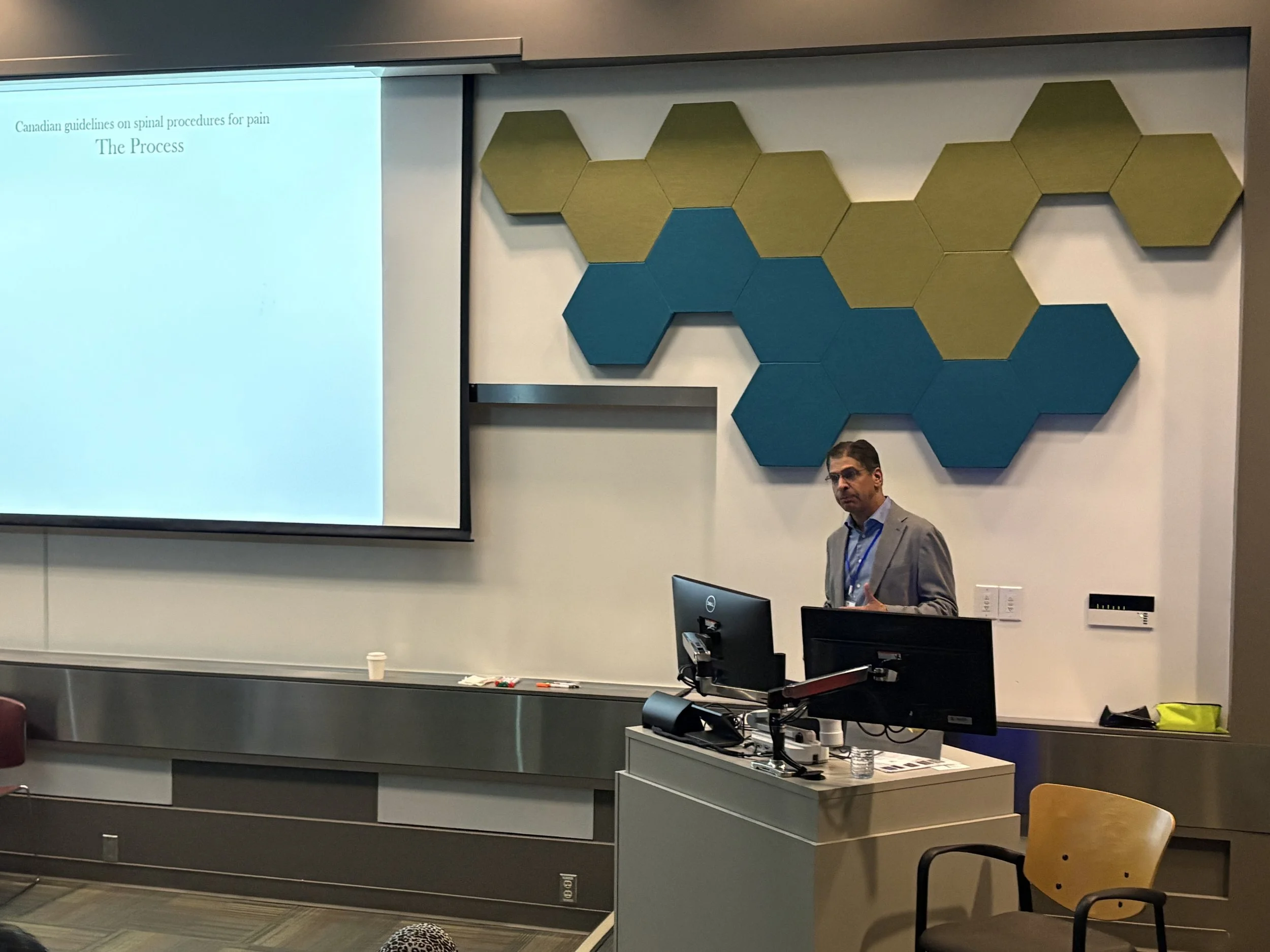 A man giving a presentation in front of a projection screen with notes about Canadian guidelines on spinal procedures for pain. There are decorative hexagonal wall panels in green and blue, and a computer on a desk with office supplies and water bott