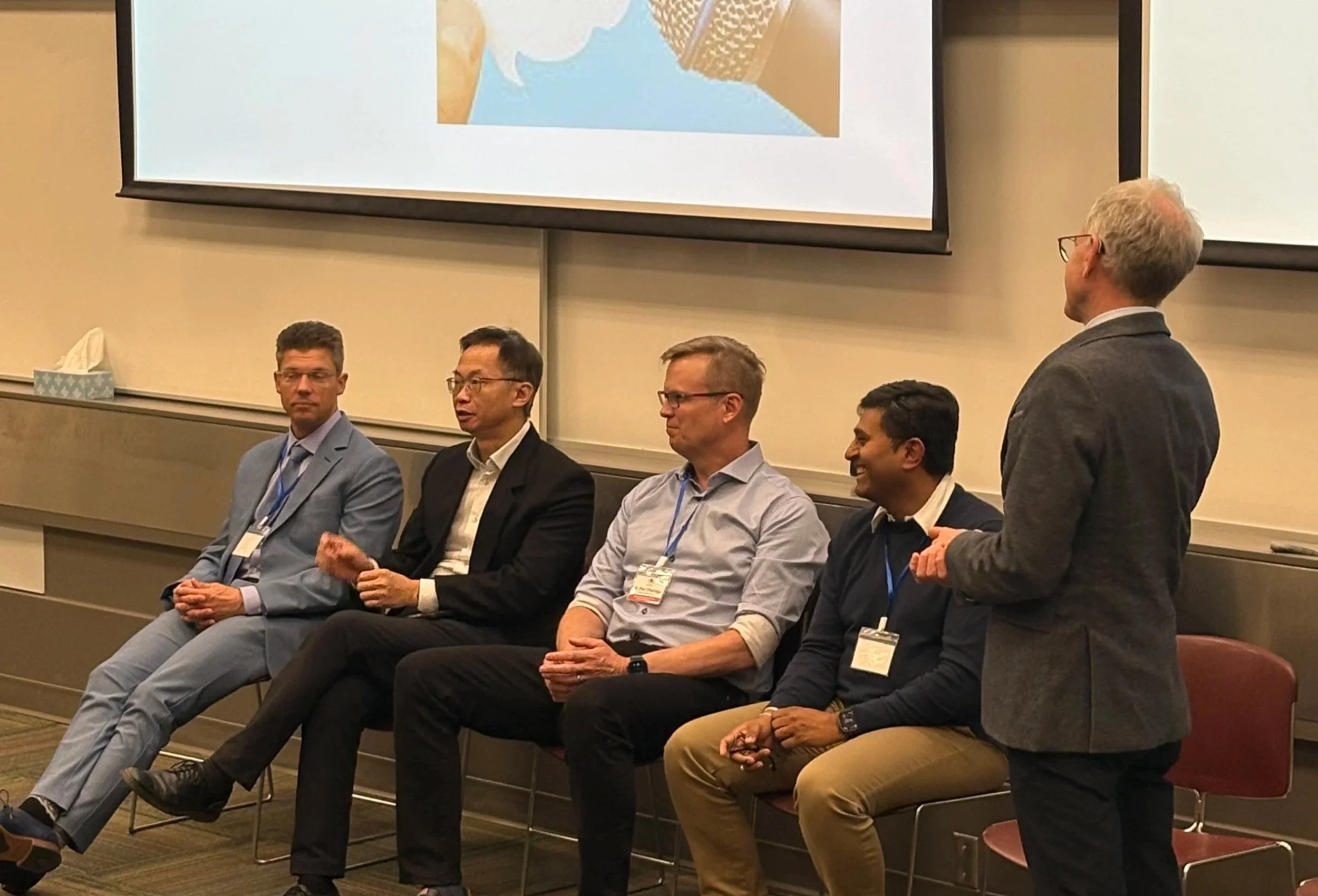 A group of five men seated in a row at a conference, with a sixth man standing and speaking to them. The men are dressed in business attire, wearing suits and dress shirts, with name tags around their necks. A large screen is visible behind them, sho