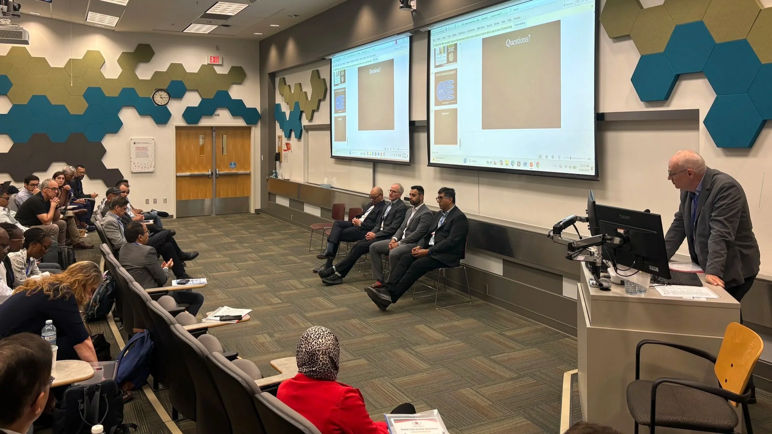 A classroom lecture hall with an audience and a panel of six men sitting near the front. A woman at a podium is speaking, and a large screen displays a presentation slide with the word "Questions?" on it.
