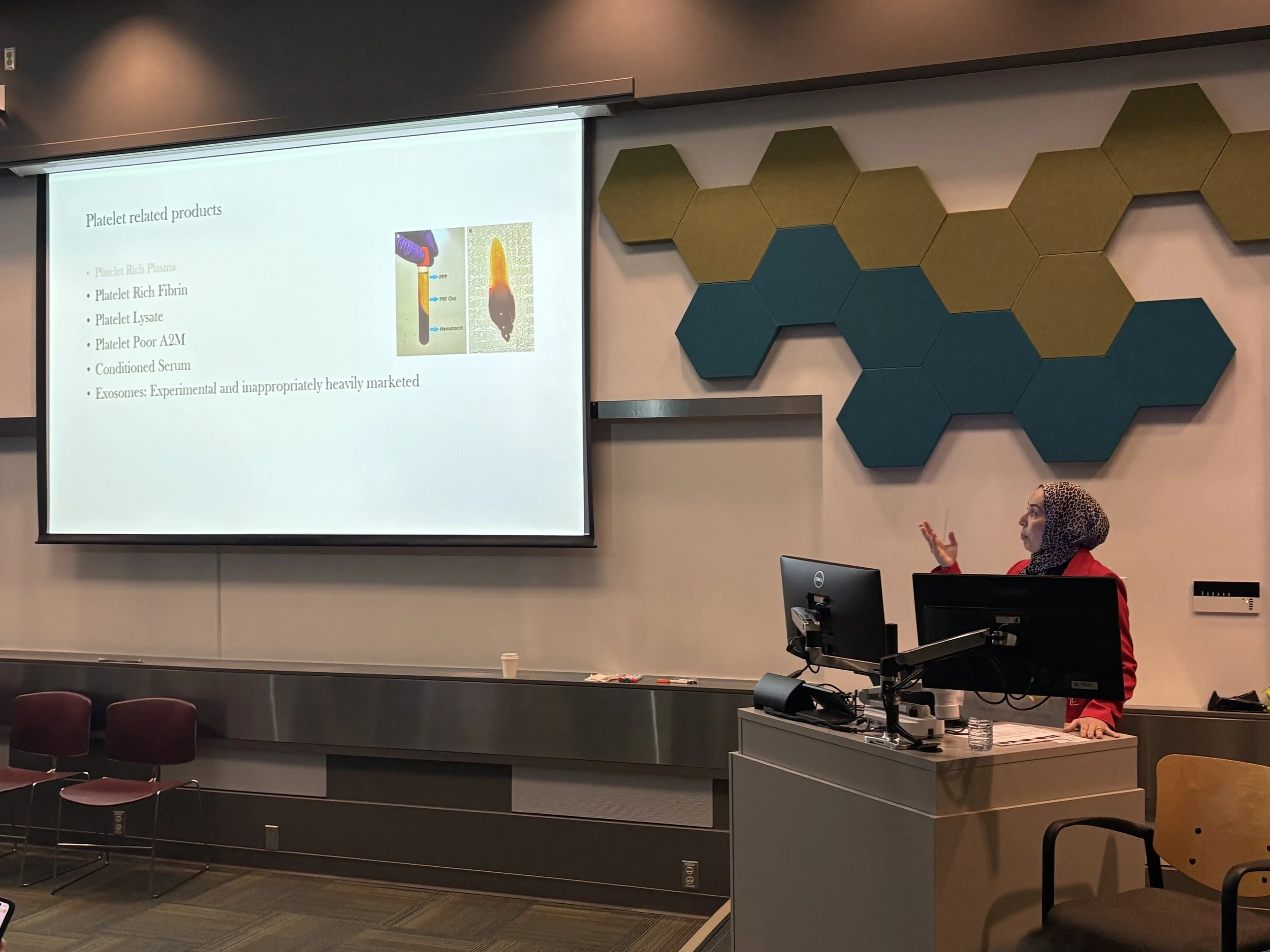 A woman in a red blazer with a leopard print headscarf giving a presentation in a conference room. She is standing at a lectern with computer monitors, and a projection screen displays a slide titled 'Platelet related products' with bullet points and
