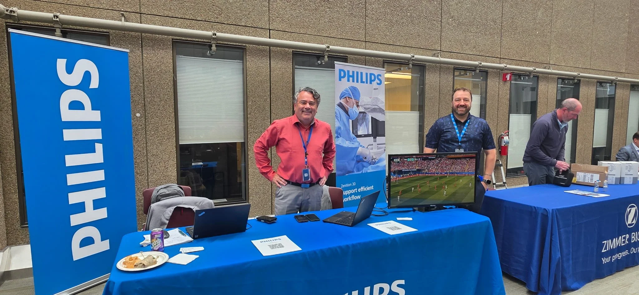 A group of four men at a Philips booth during an event, with displays including laptops, a monitor showing a sports game, and Philips banners.