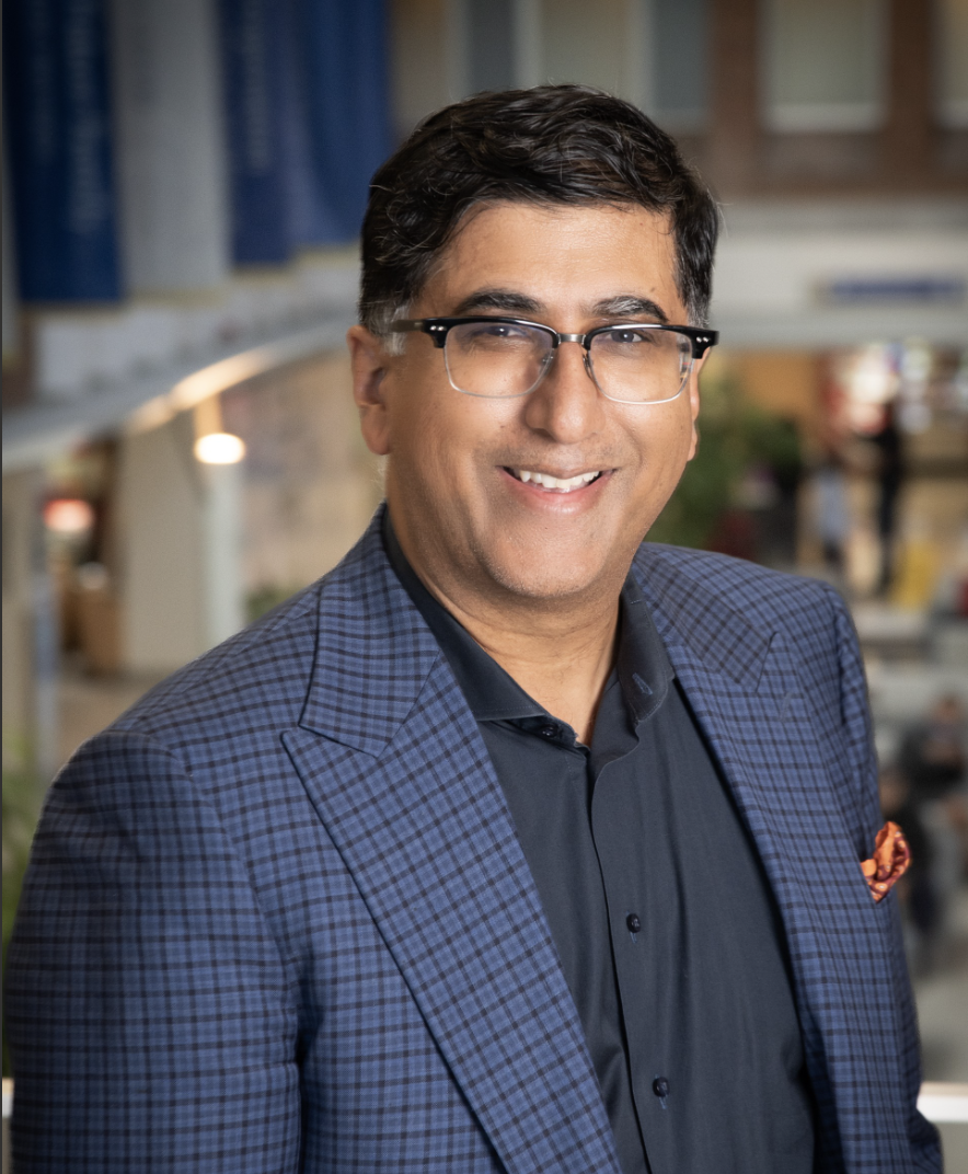A man with dark hair and glasses smiling, wearing a dark shirt and a checkered blazer, in an indoor setting