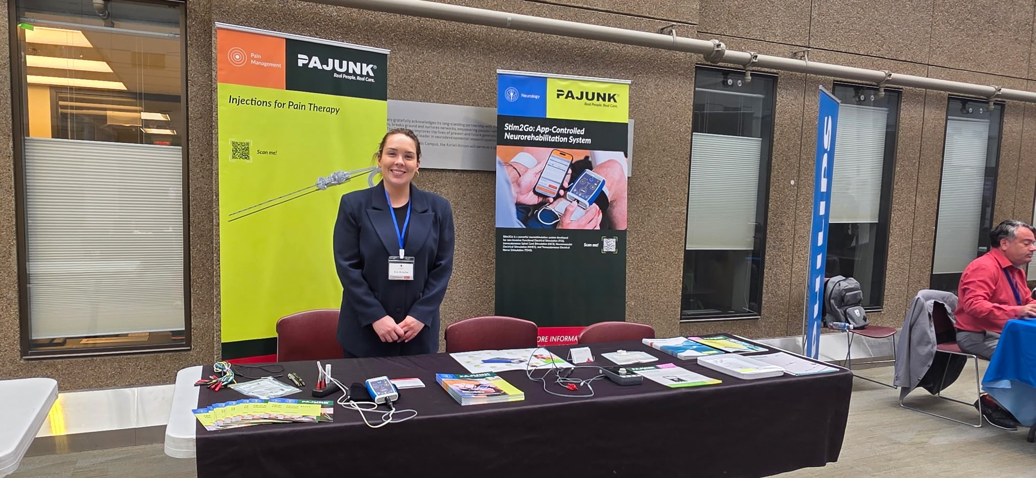 A woman standing behind a table at a health or medical conference booth with banners displaying information about pain management and neurorehabilitation systems, with informational pamphlets and electronic devices on the table.