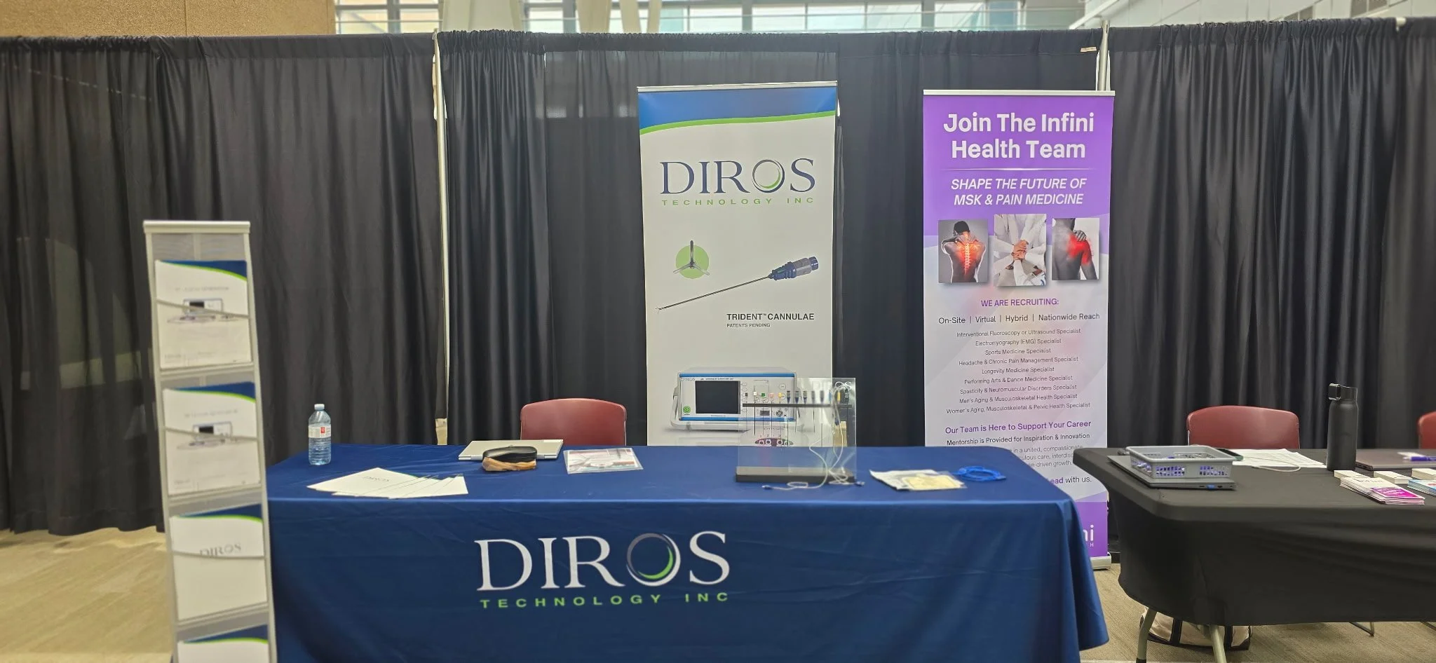 An exhibition booth with banners displaying DIROS Technology Inc., and information about joining the Infini Heath Team for medical and pain management solutions, featuring support staff and promotional materials.