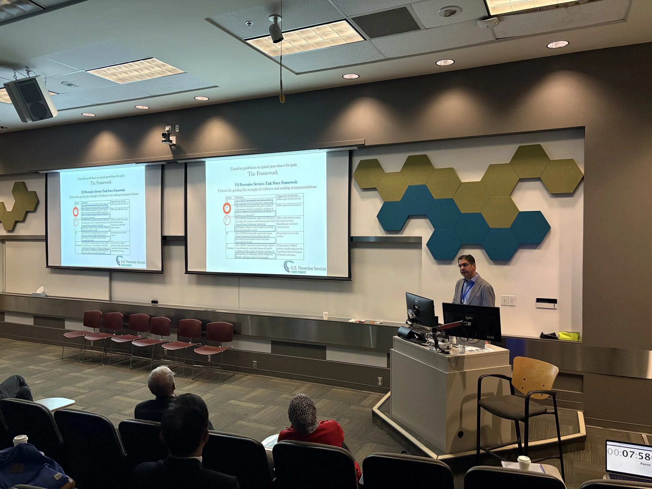 A lecture hall or conference room with a presenter standing near a podium and audience members seated listening. Two screens display presentation slides with text and graphics. The ceiling has recessed lighting and speakers, and there are decorative 