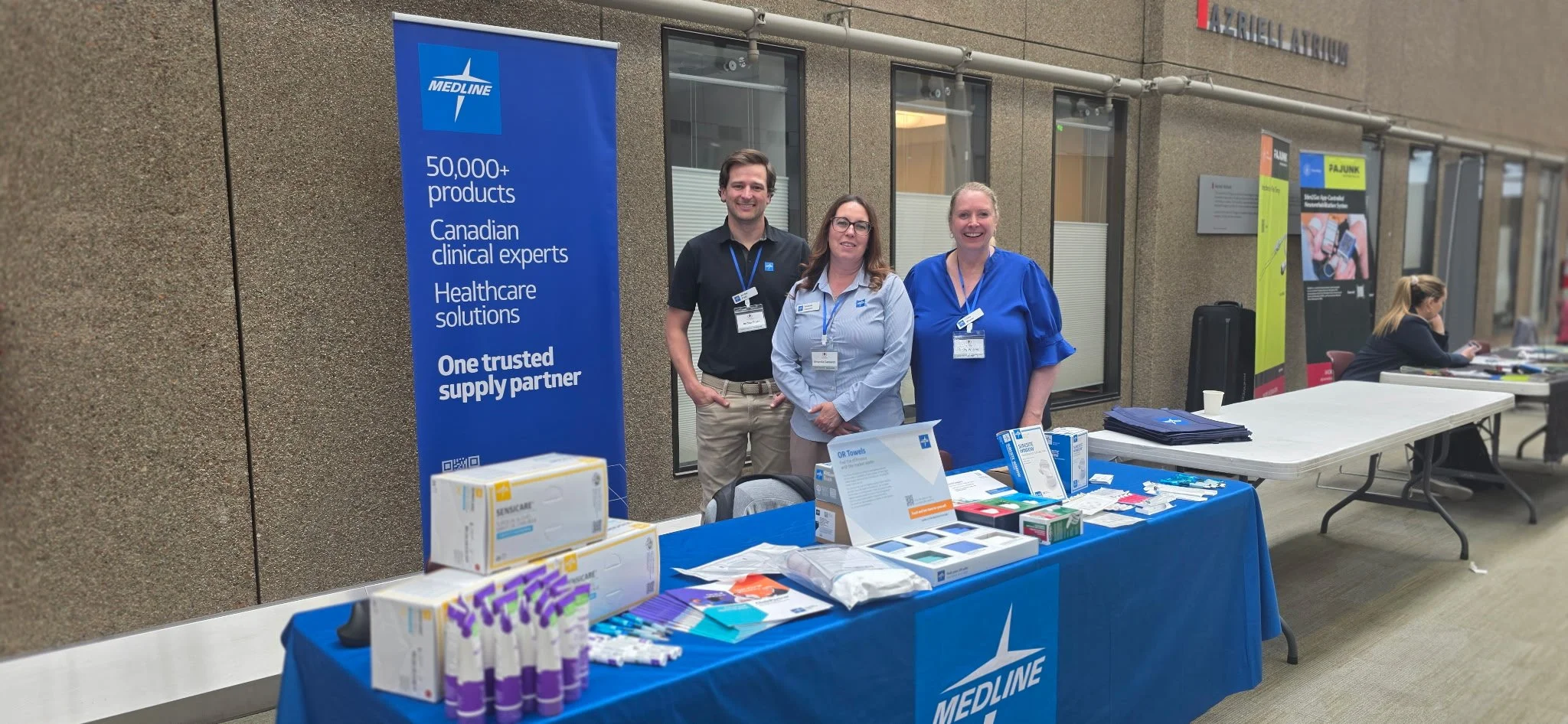 Three people standing behind a table at an event booth for Medline, displaying medical supplies. The booth has a blue tablecloth and a large blue banner with white text. There are various medical products on the table, and a woman seated at a nearby 