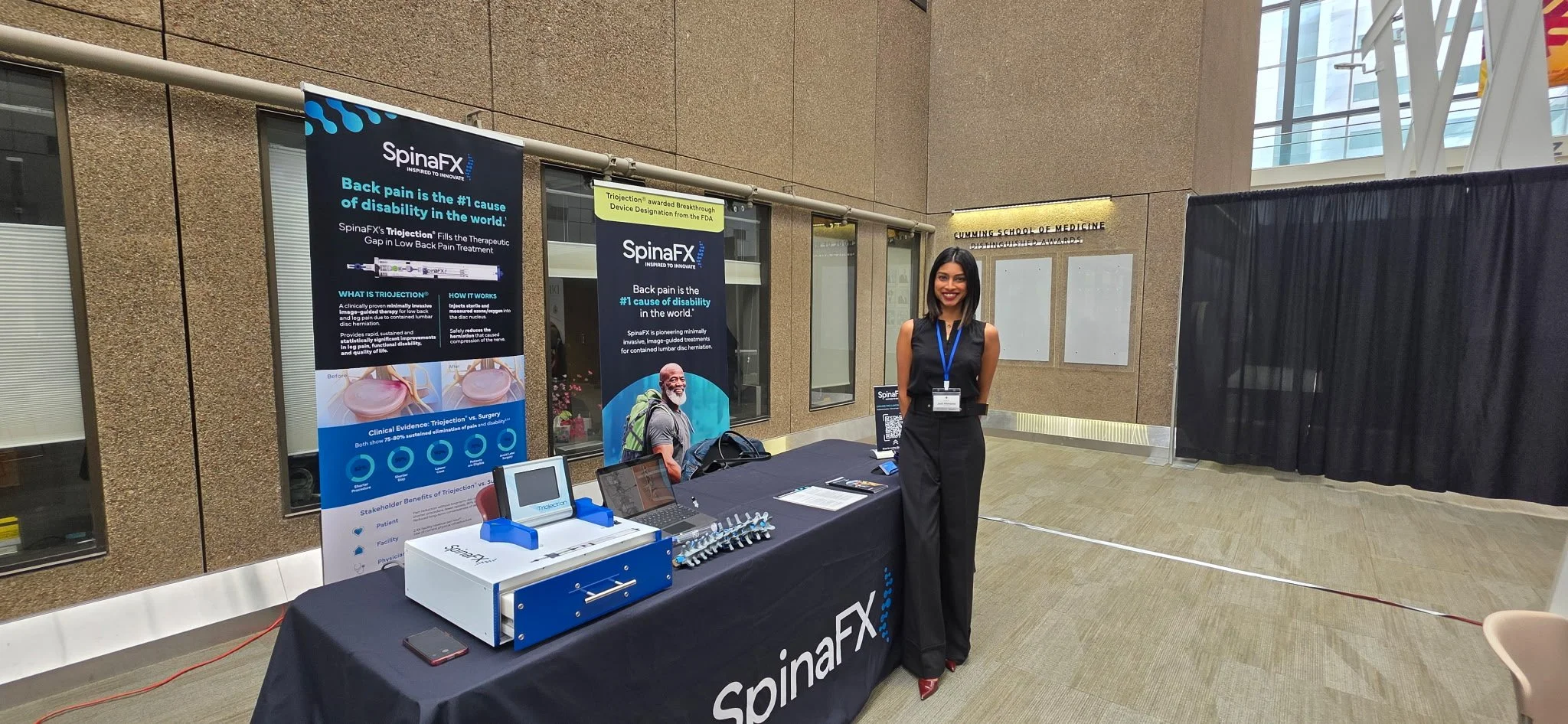 A woman in formal attire standing next to a table with promotional materials at an exhibition booth for SpinaFX, which features banners about back pain treatment and a device called Triojection. The booth is set up in a modern, indoor space near a do