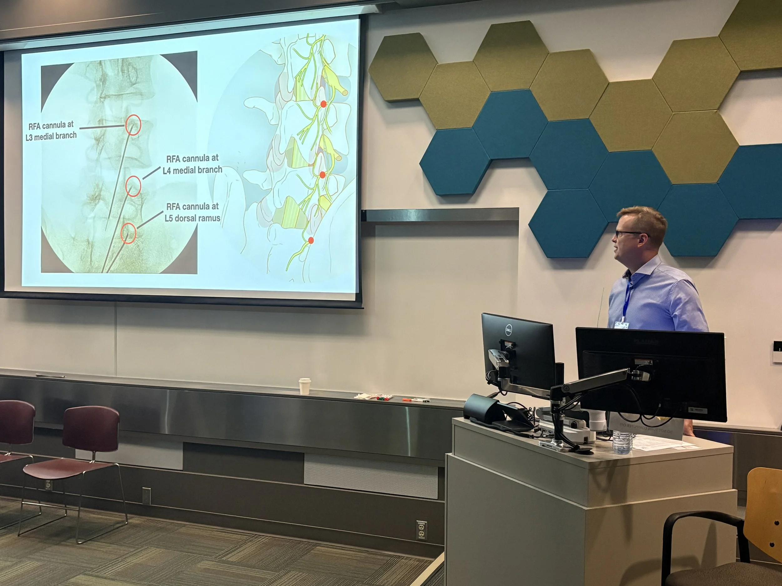 A classroom or seminar room with a presentation slide projected on the front wall showing diagrams of the nervous system and labels for specific anatomical parts. A man in a light blue shirt stands near a computer at the front of the room, and there 