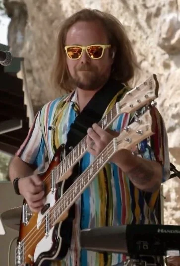 Man with shoulder-length hair and a beard playing multiple guitars, wearing striped colorful shirt and yellow sunglasses, in front of a rocky background.