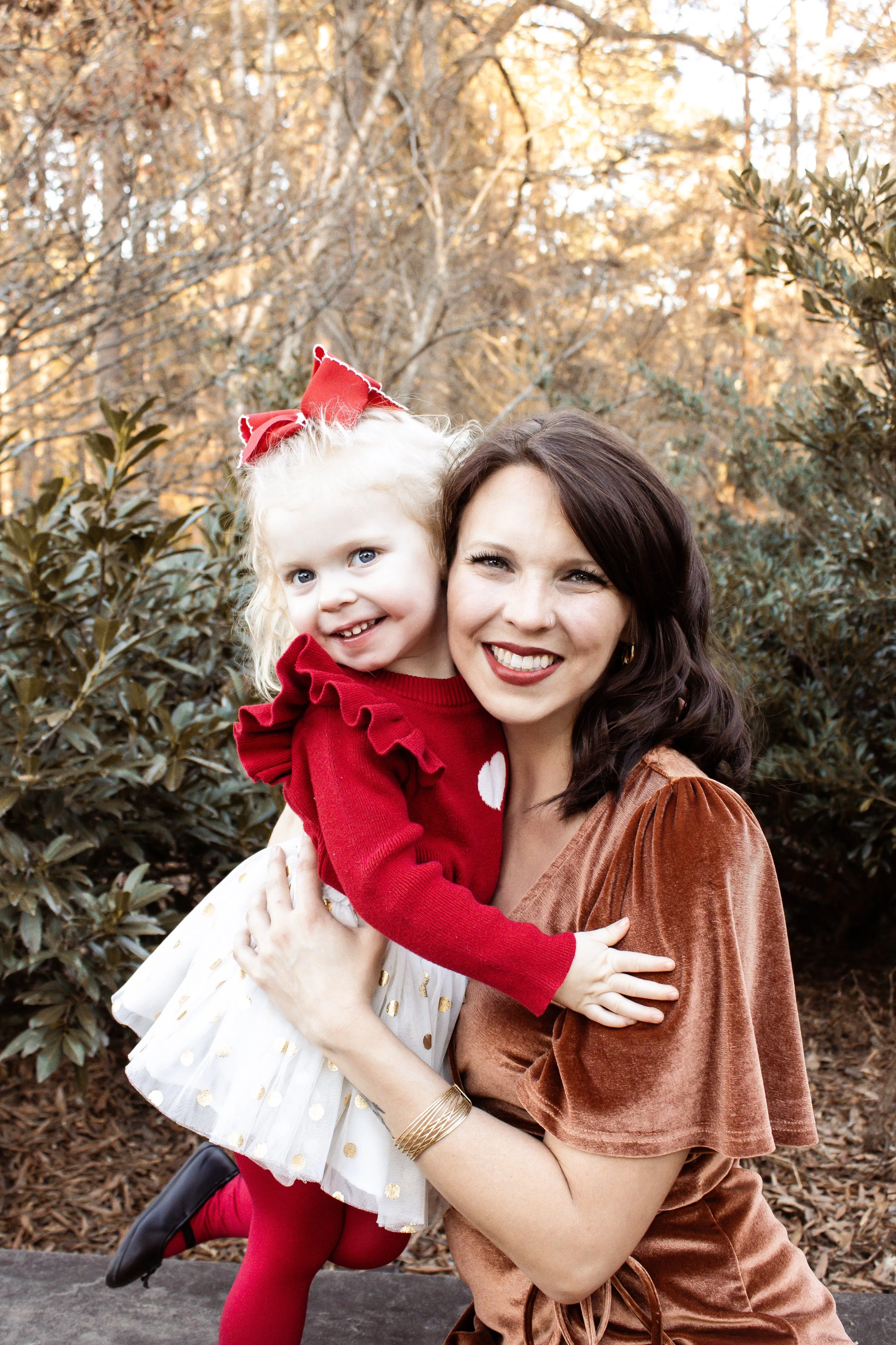 A woman and a young girl smiling and hugging outdoors during autumn, surrounded by trees and bushes.
