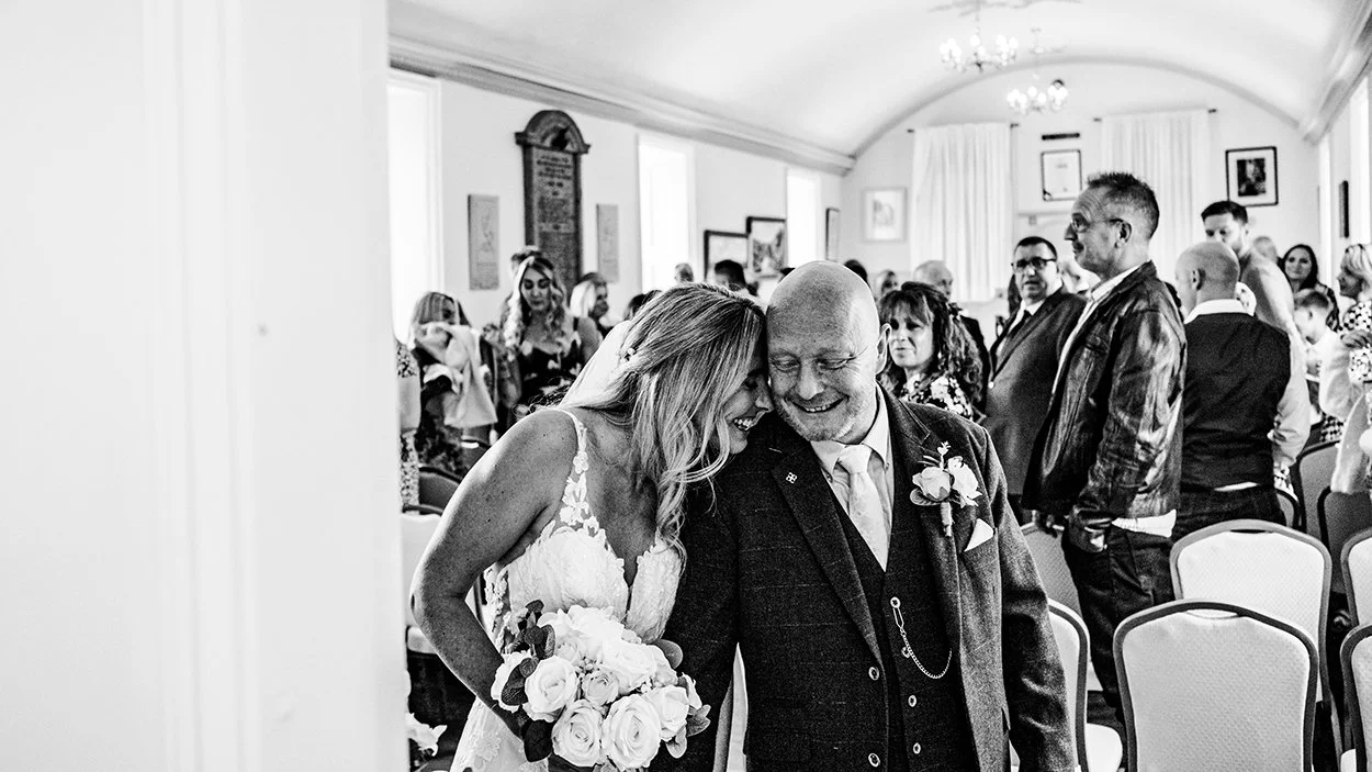 A bride and an older man smiling and leaning head to head during a wedding ceremony, with guests in the background, taken by Oliver Harris, photographer, unposed photography.