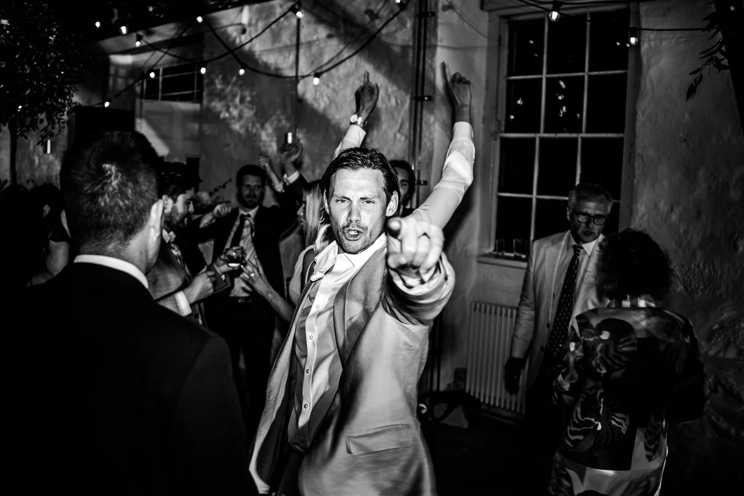 A group of people dancing and socializing at a party or celebration in a dimly lit room, with string lights overhead, black and white photo, with a man in a light-colored suit pointing at the camera.