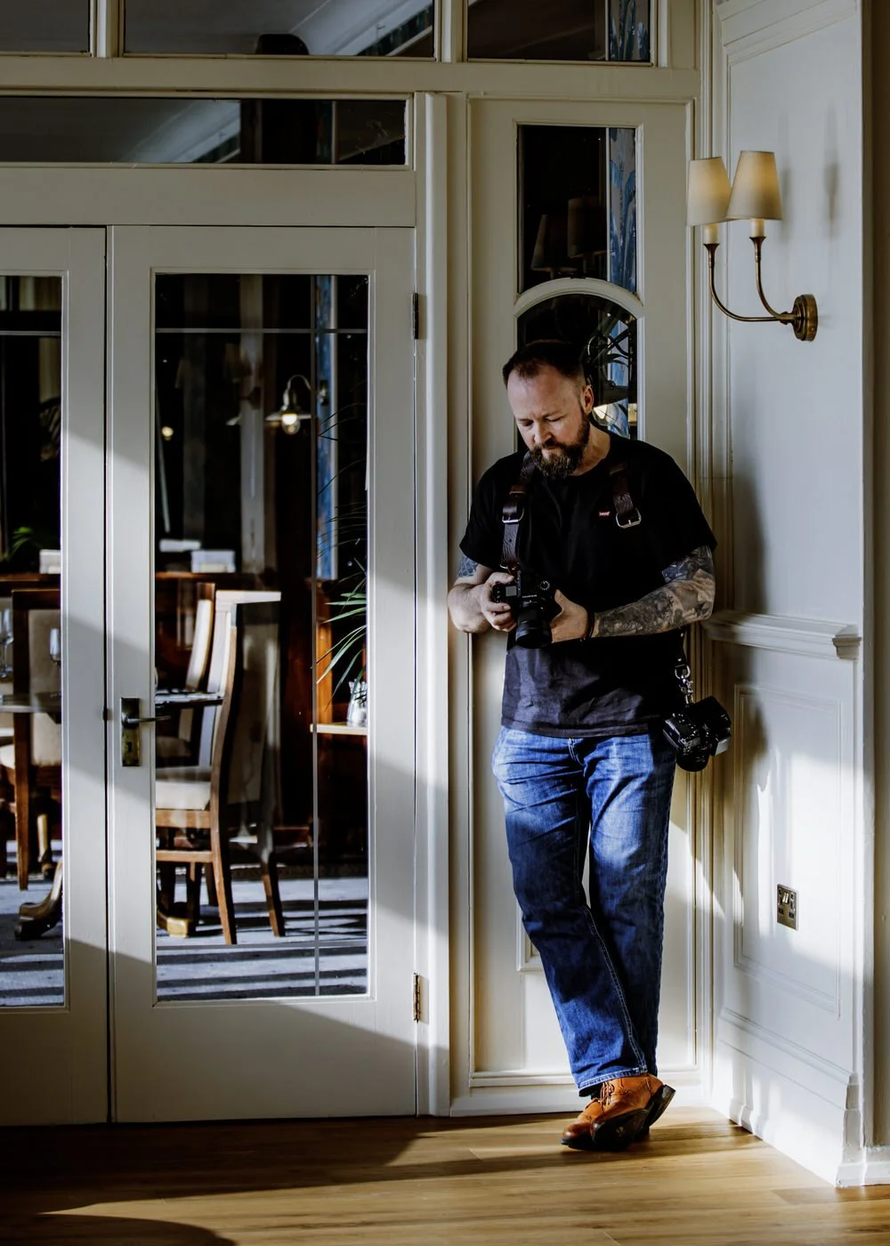 A man with tattoos, wearing a black t-shirt and blue jeans, is standing indoors, leaning against a wall, looking at a camera he is holding. He has another camera hanging from his neck. The room has cream-colored walls, a wall-mounted lamp with two shades, and glass-paneled doors revealing a dining area with chairs and a table in the background.