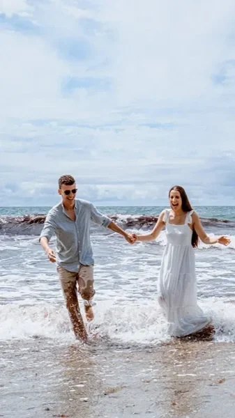 A happy couple holding hands and walking in the ocean waves at the beach on a cloudy day, taken by Oliver Harris, photographer, unposed photography.