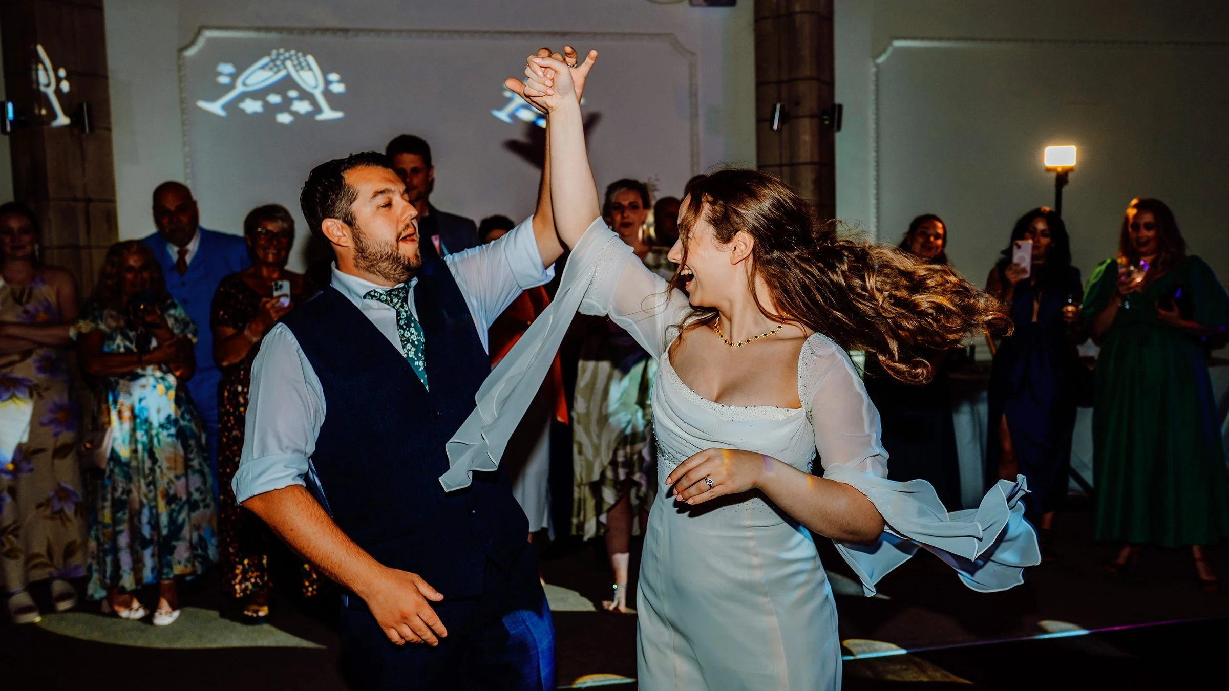 A dynamic candid shot by Oliver Harris Wedding Photographer in Cornwall captures a bride in a white flowing dress and groom in a waistcoat laughing while twirling during their energetic wedding reception dance, surrounded by cheering guests.