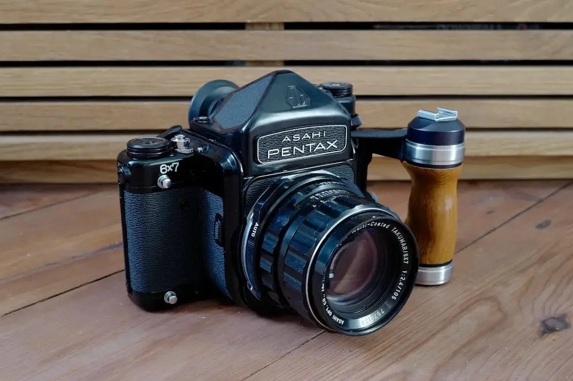 A vintage Asahi Pentax film camera with a wide lens and a wooden-handled grip, placed on a wooden surface with a wooden slat background.