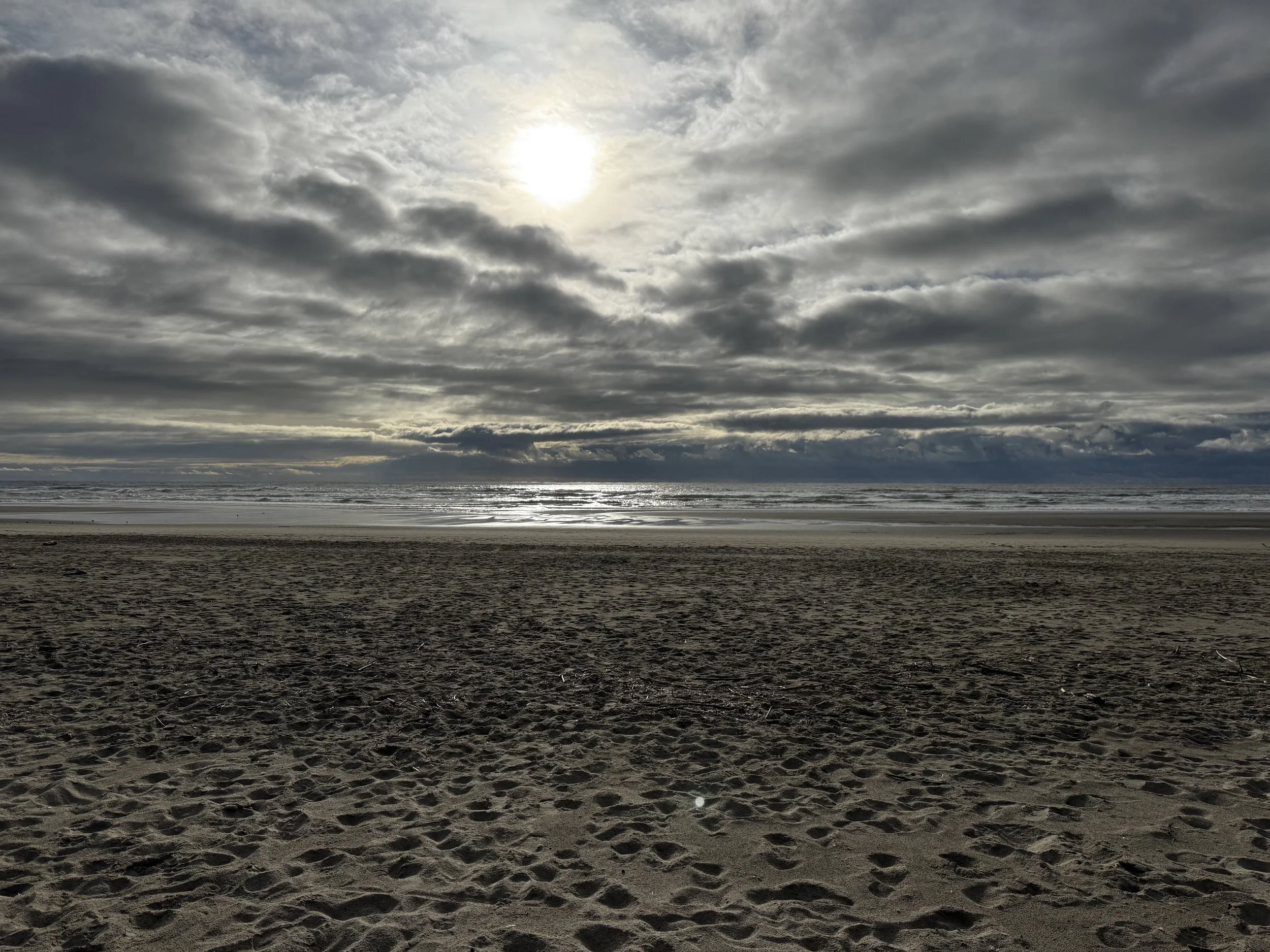Cloudy beach with the sun visible through the clouds, ocean waves, and sandy shore in the foreground.
