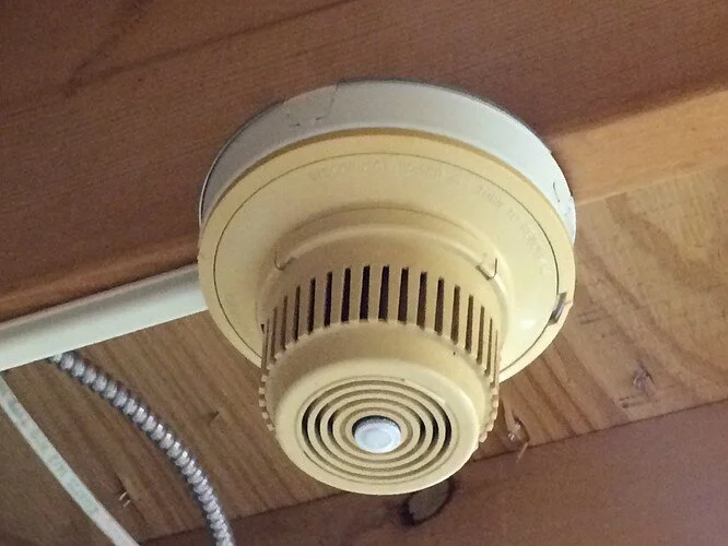 A ceiling-mounted smoke detector on a wooden ceiling with exposed wiring and a metal conduit.