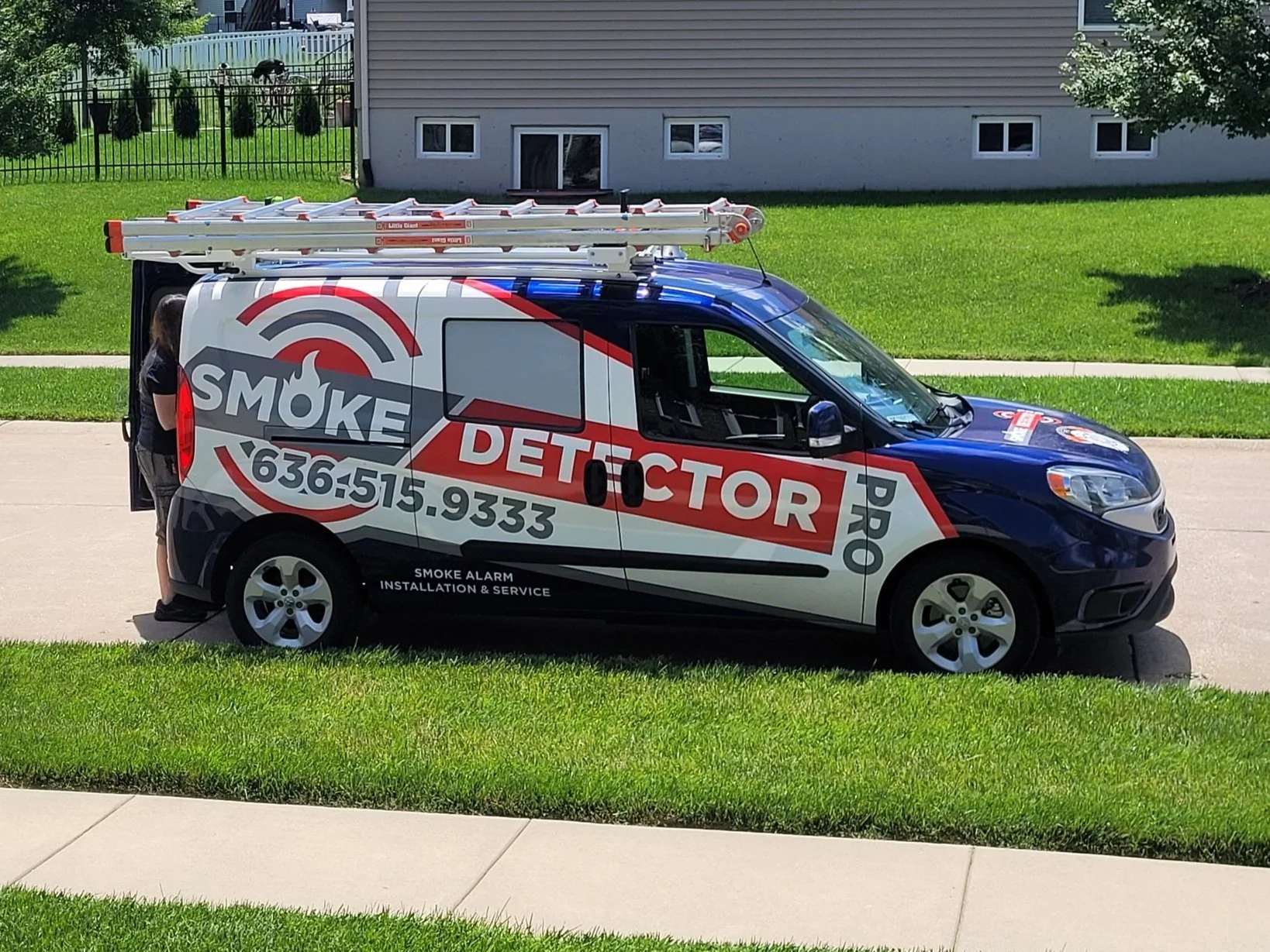 Commercial van with "Smoke Detector Pro" branding and contact information, parked on a residential street, featuring a ladder on the roof.