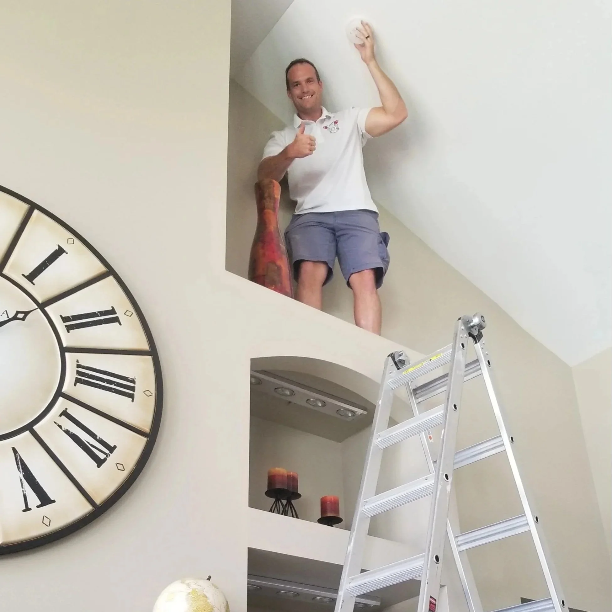 A man standing on a ladder, giving a thumbs-up and smiling while holding a smoke detector on the ceiling in St. Louis MO
