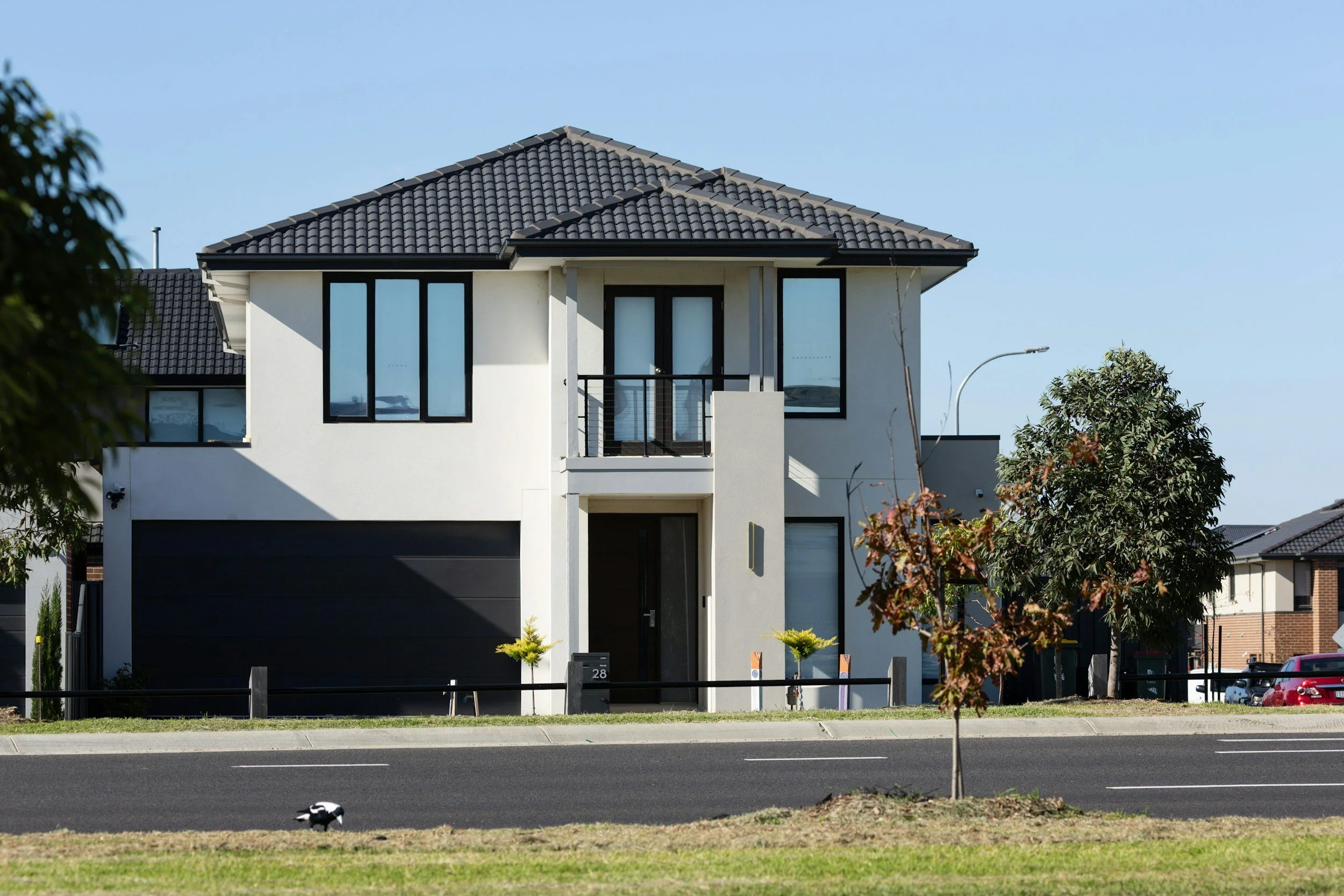 Modern two-story house with a black tiled roof, white walls, and large windows, situated along a street with trees and parked cars.