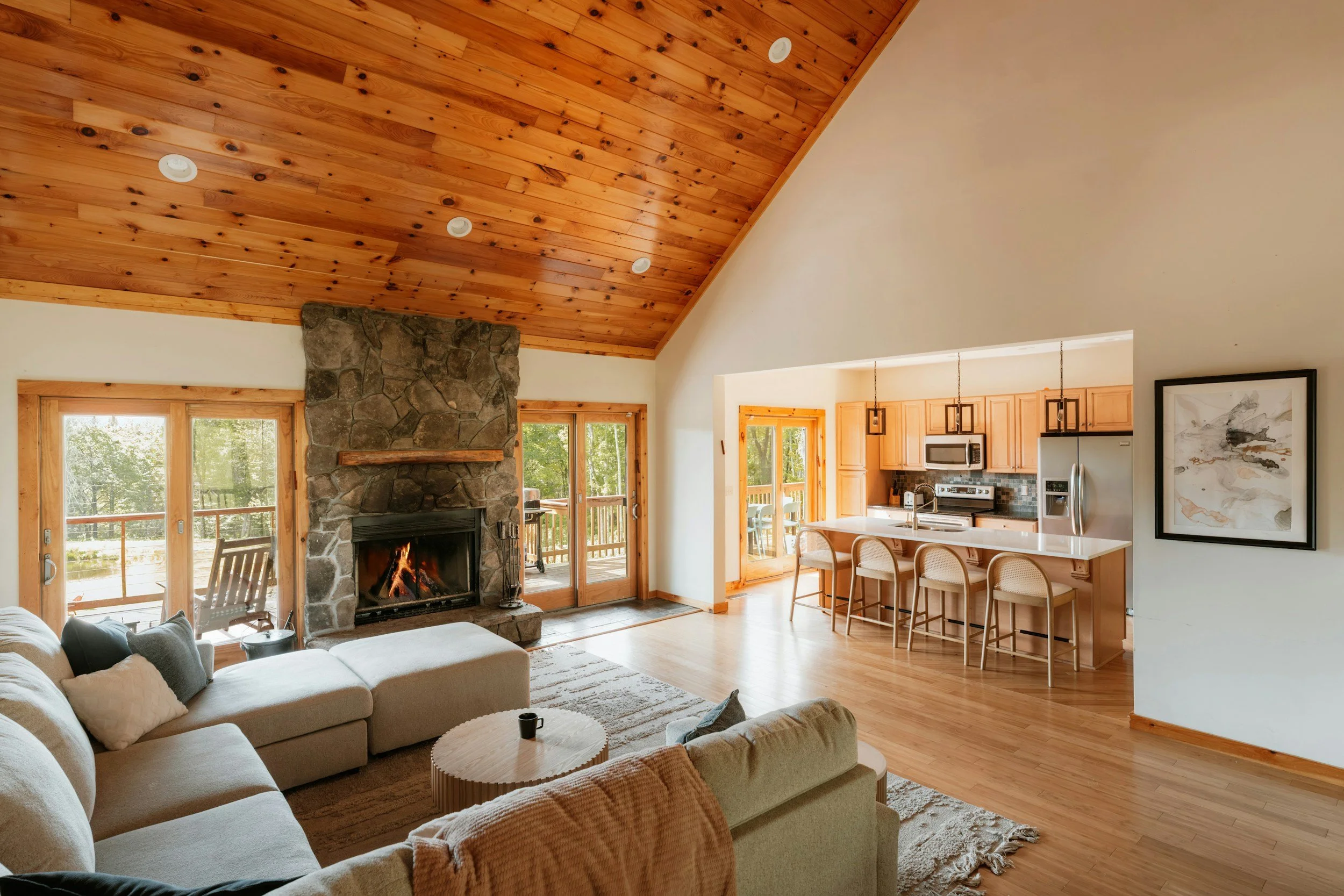 A living room with a wooden vaulted ceiling, stone fireplace with fire burning, large sliding glass doors showing trees outside, and a beige sectional sofa with pillows. Adjacent is a kitchen with light wood cabinets, a white countertop, pendant lighting, and a framed artwork.
