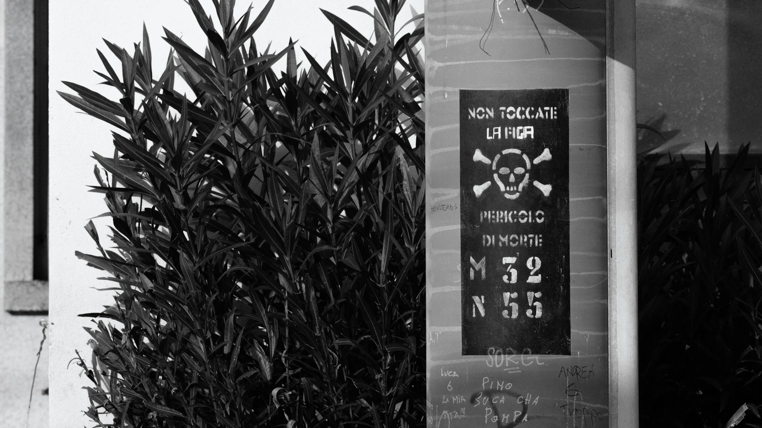 A black and white photo of a sign with a skull and crossbones symbol that reads 'NON TOCATE LA PIZZA, PERICOLO DI MORTE' and a list of names below.
