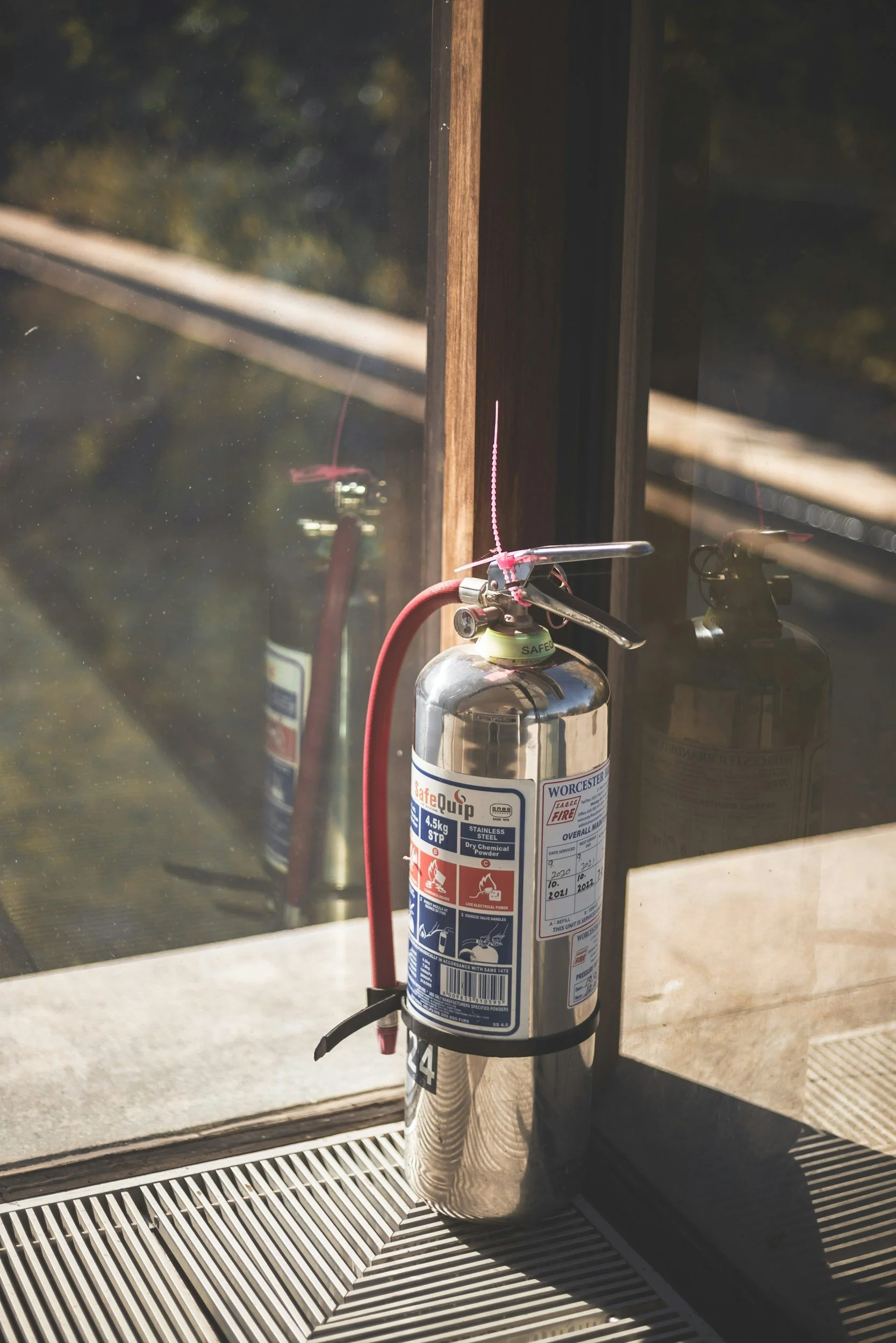 A fire extinguisher mounted near a window, with a pink electrical wire attached to its handle, in a building interior.