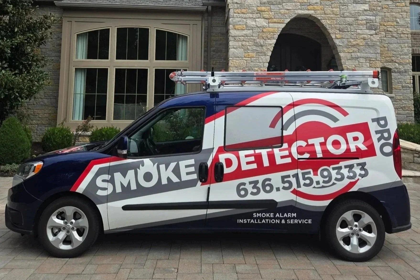 A van parked in front of a house with gray stone walls and a large window. The van has a red, white, and gray design and is labeled 'Smoke Detector Pro' with a phone number and services related to smoke alarm installation and service. A ladder is secured on top of the van.