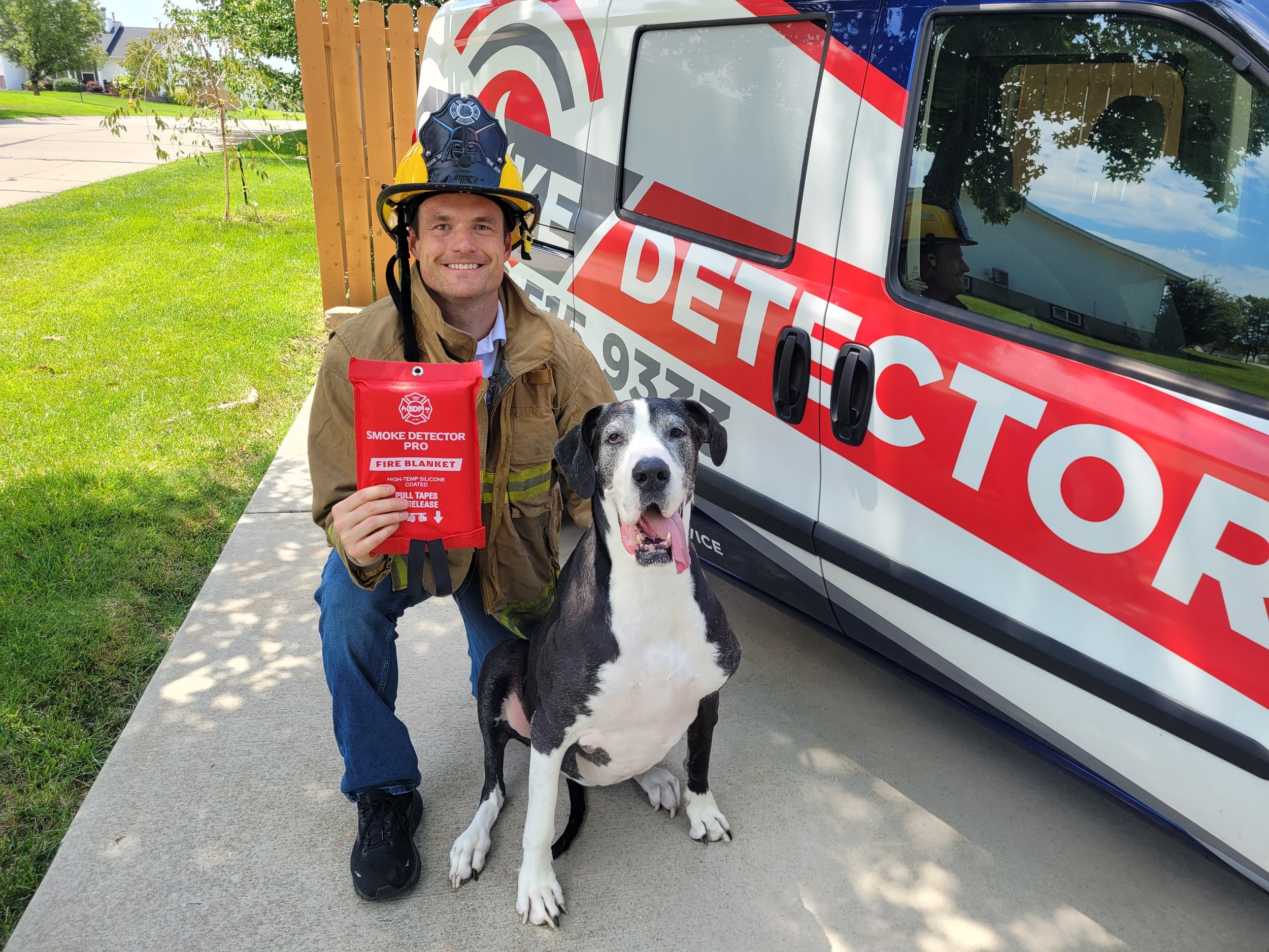 Firefighter with helmet and fire blanket kneeling next to a Great Dane in front of a smoke detector service van.
