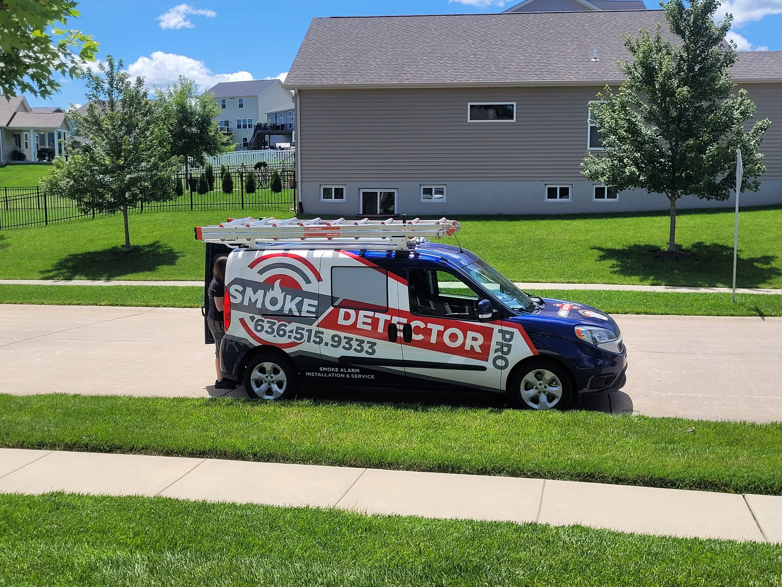 A black and white service van with red and gray graphics, labeled 'Smoke Detector PRO,' is parked on a driveway in front of a green lawn and houses. The van has ladders on top and contact information printed on the side.