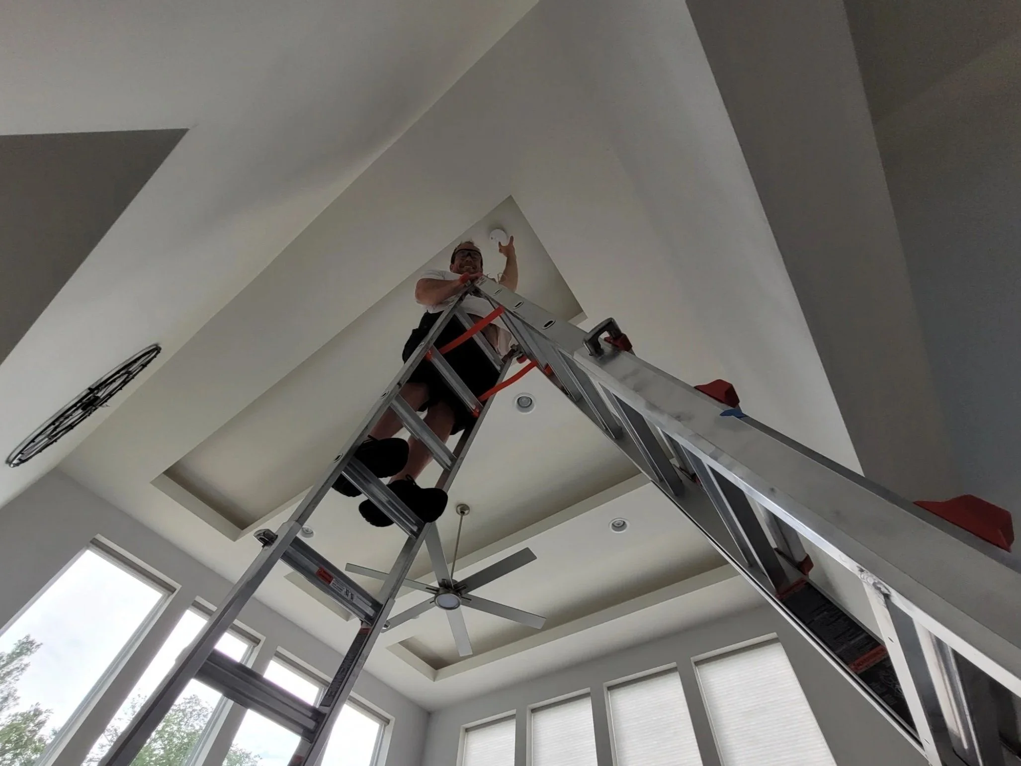 A person standing on a ladder reaching up towards the ceiling in a modern room with large windows and a ceiling fan.