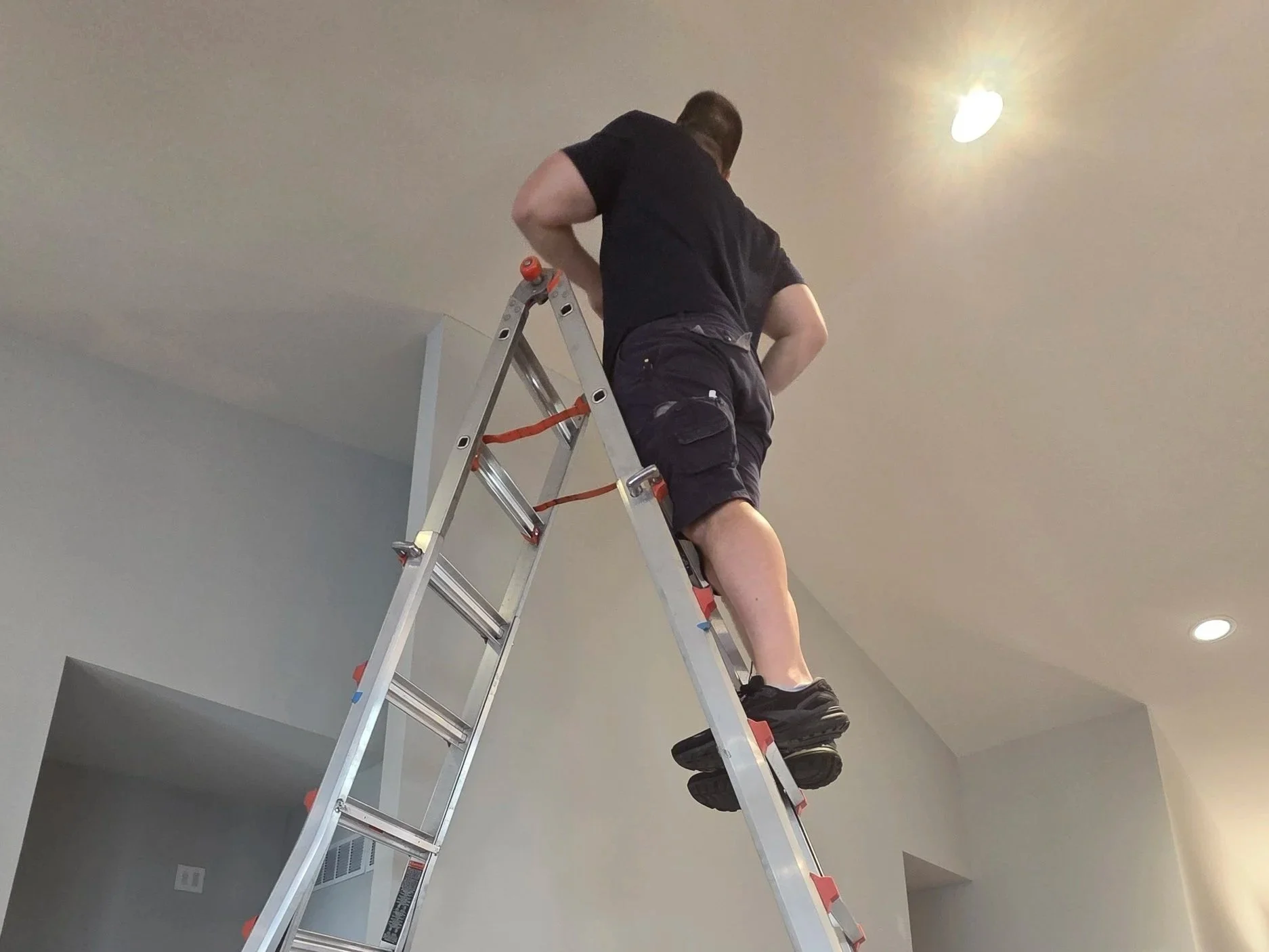 Person standing on a step ladder working on the ceiling in a room with light-colored walls and ceiling lights.